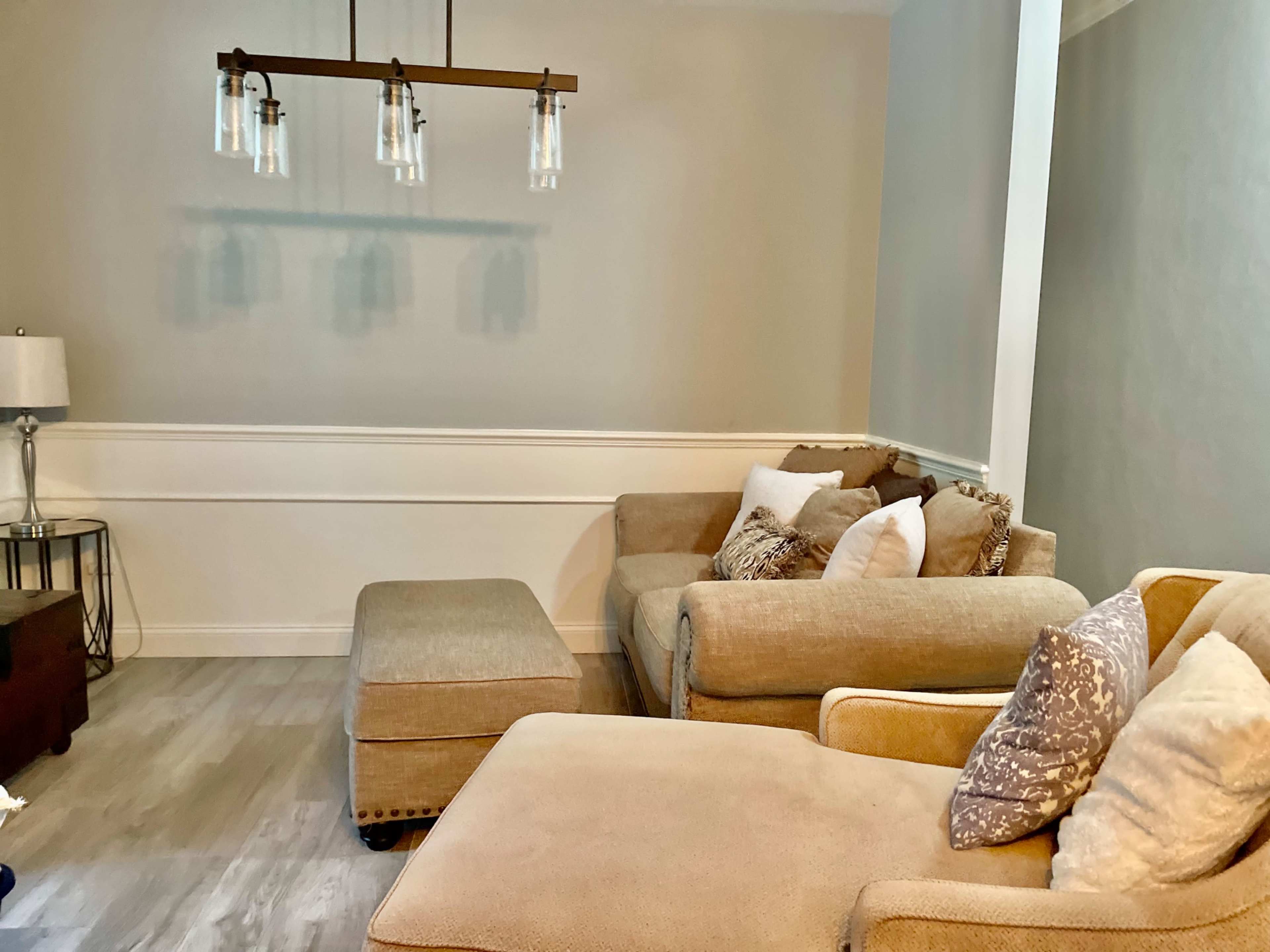 A light-colored living room with a sectional sofa, an ottoman, and a modern light fixture hanging from the ceiling.