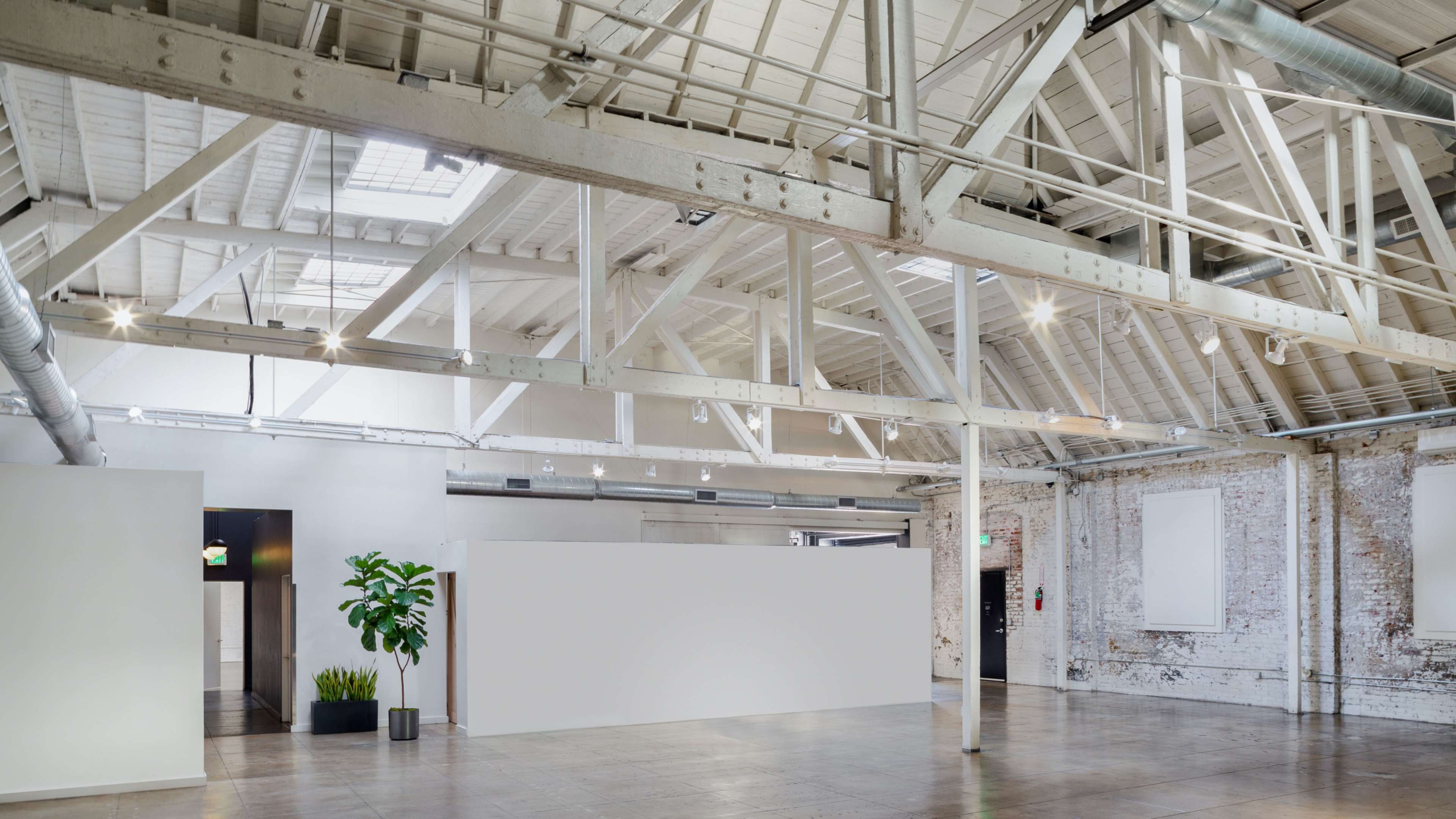 The image shows a spacious, industrial-style interior with wooden beams, large windows, and a white partition surrounded by potted plants.