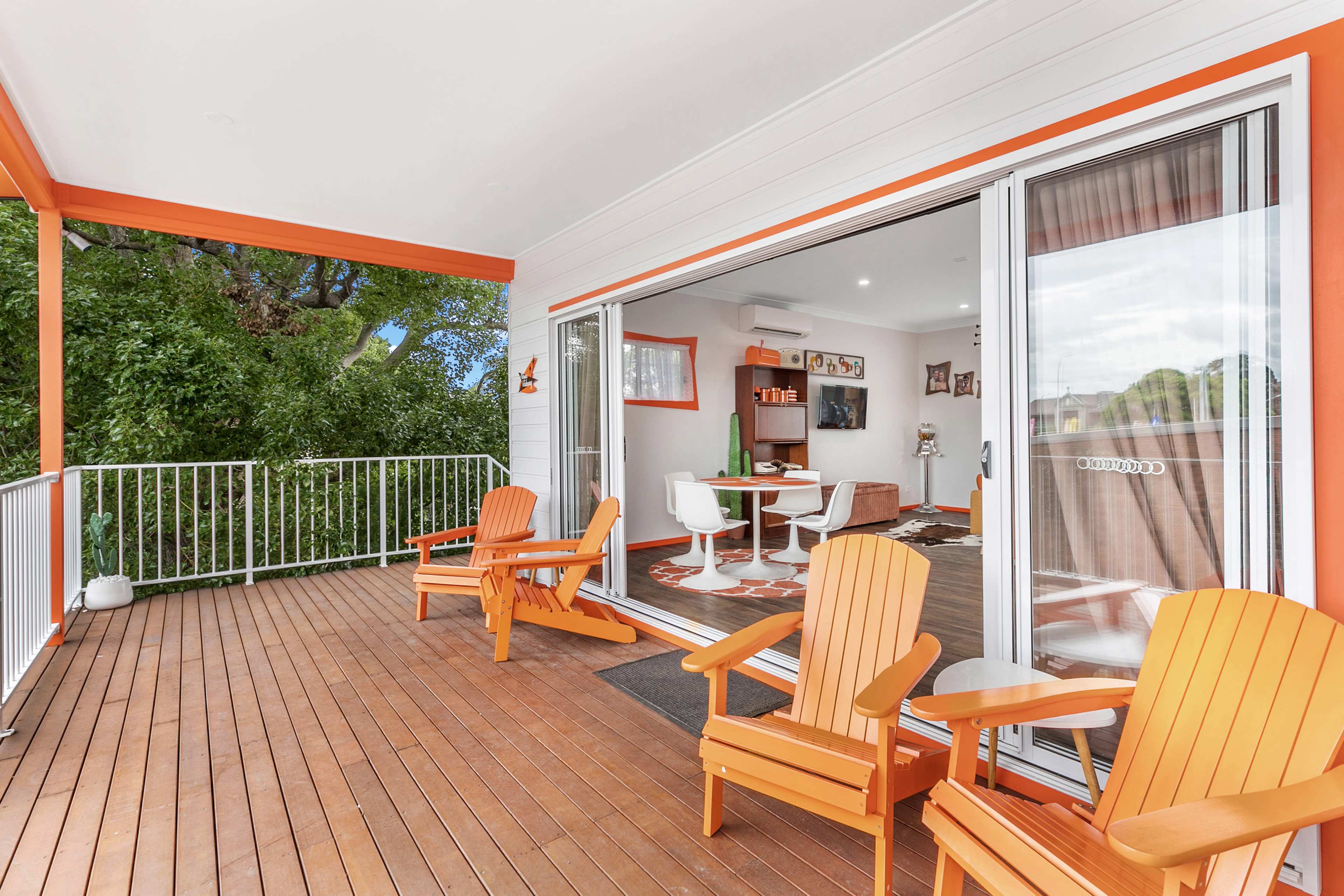 A spacious balcony features orange Adirondack chairs overlooking a green area, with sliding doors leading to a modern living space inside.