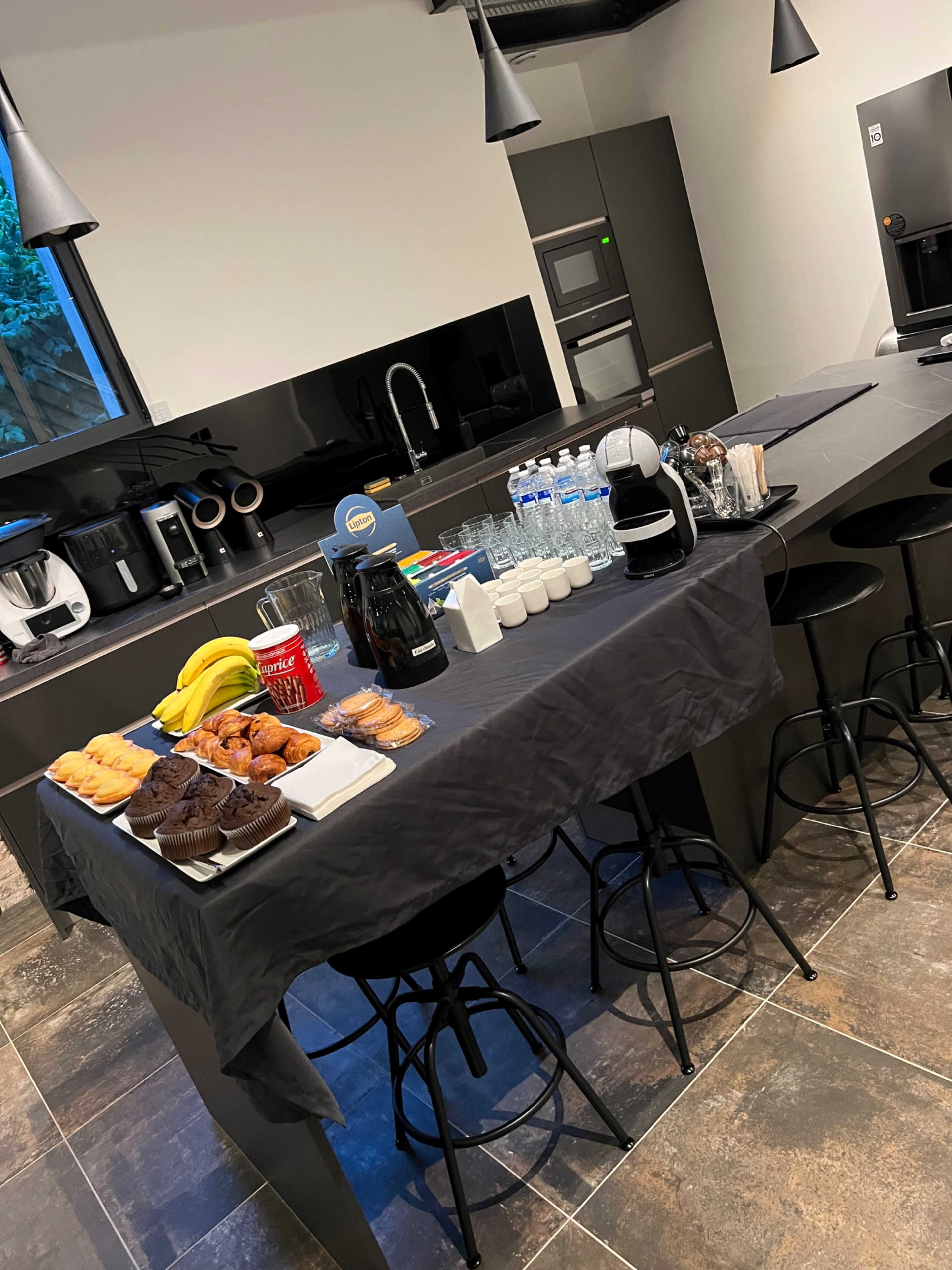 A modern kitchen counter is set up with a variety of snacks, including pastries, muffins, and bananas, alongside drinks and utensils.