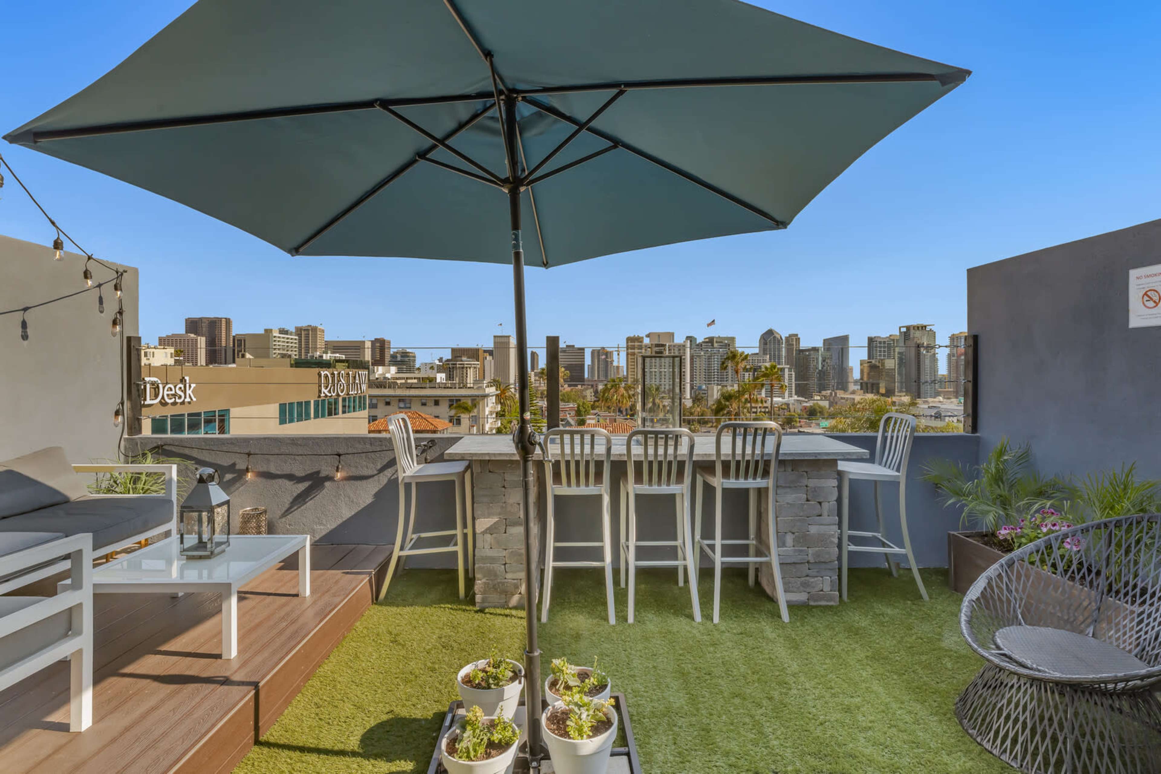A rooftop patio with an umbrella, seating, and planters, overlooking a city skyline.
