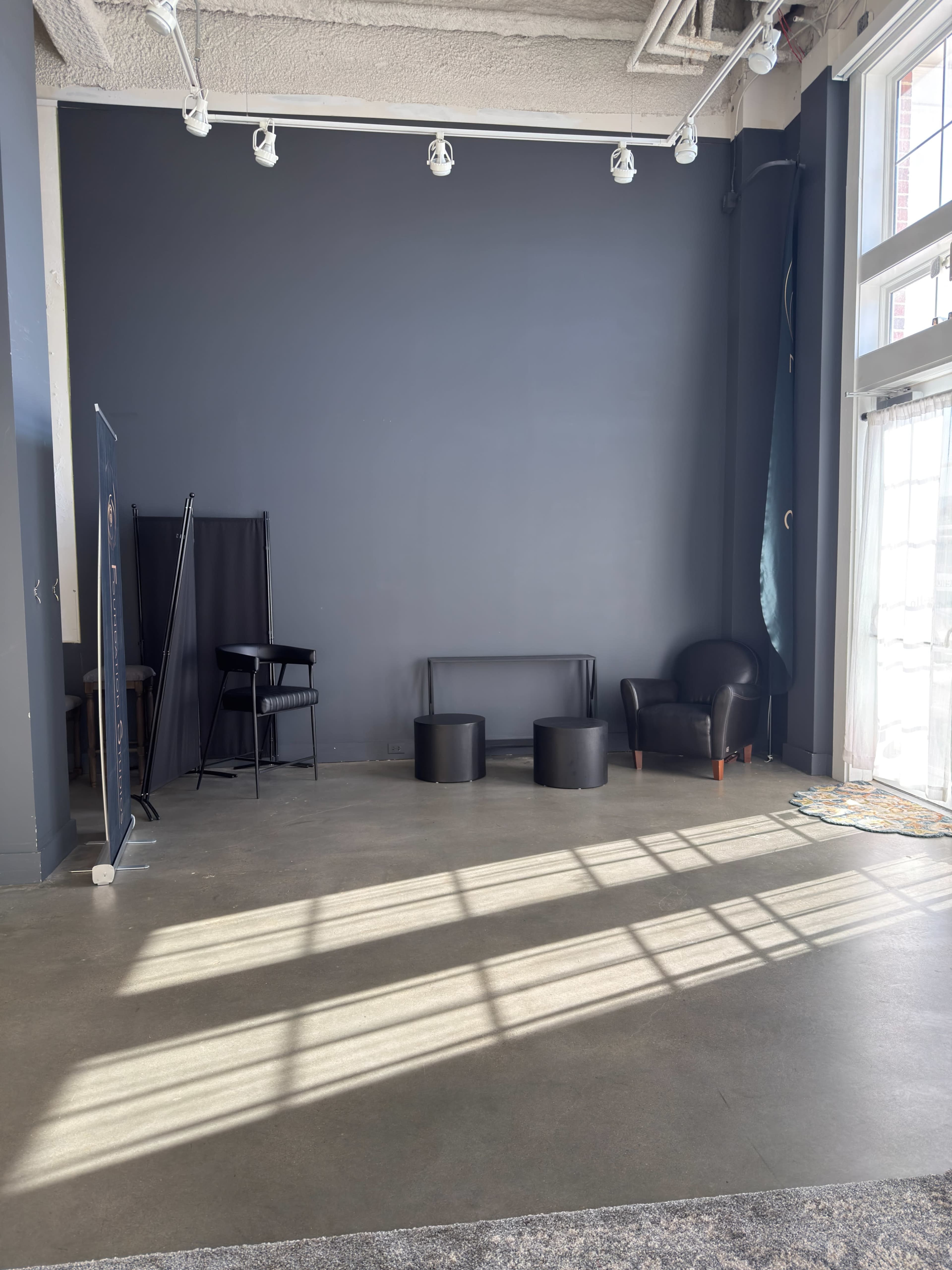 The image shows an empty interior space with a dark wall, a black chair, two cylindrical tables, and a leather armchair, illuminated by sunlight casting shadows on the floor.
