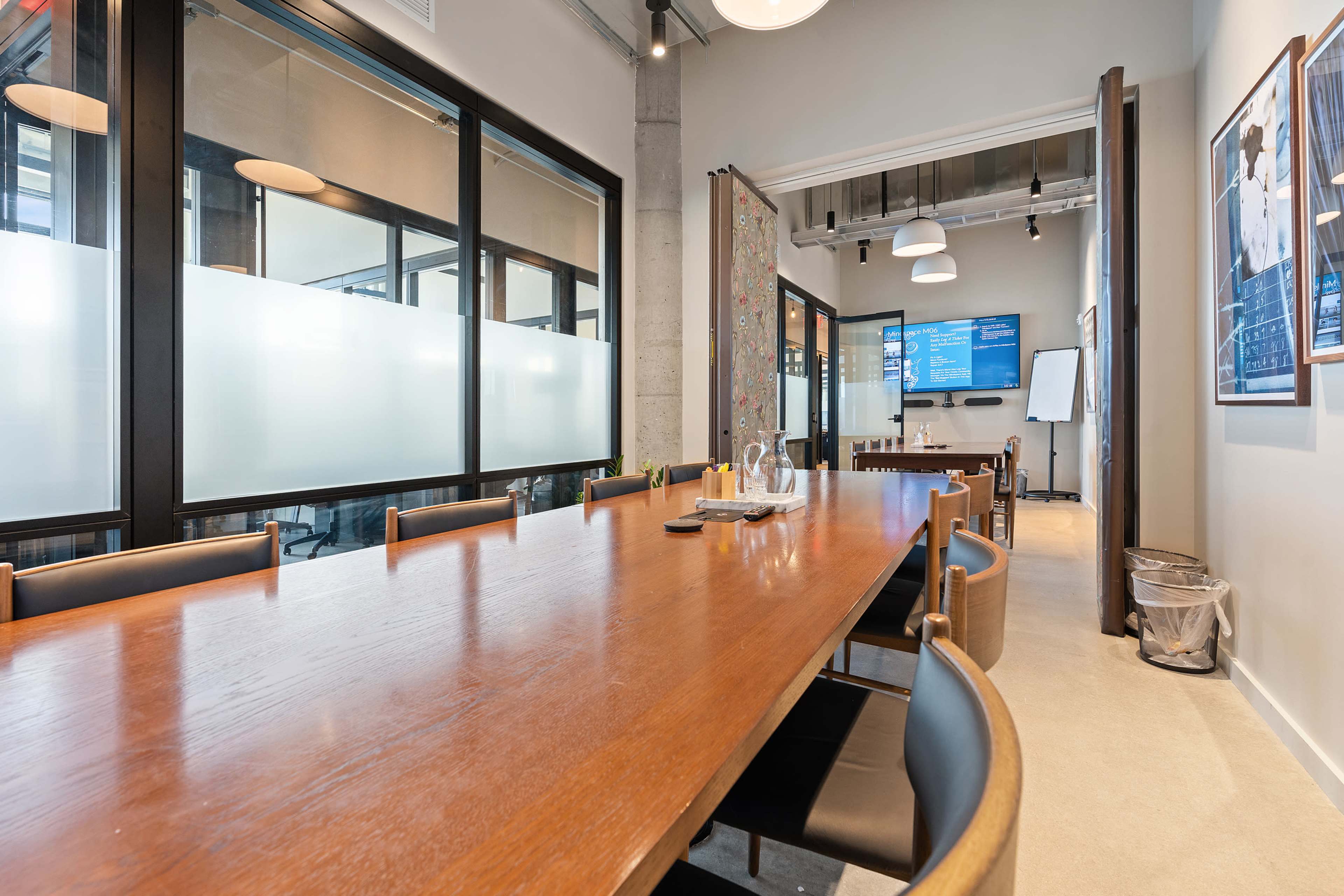 A large wooden conference table is set in a modern meeting room with glass partitions, displaying a screen and whiteboard in the background.