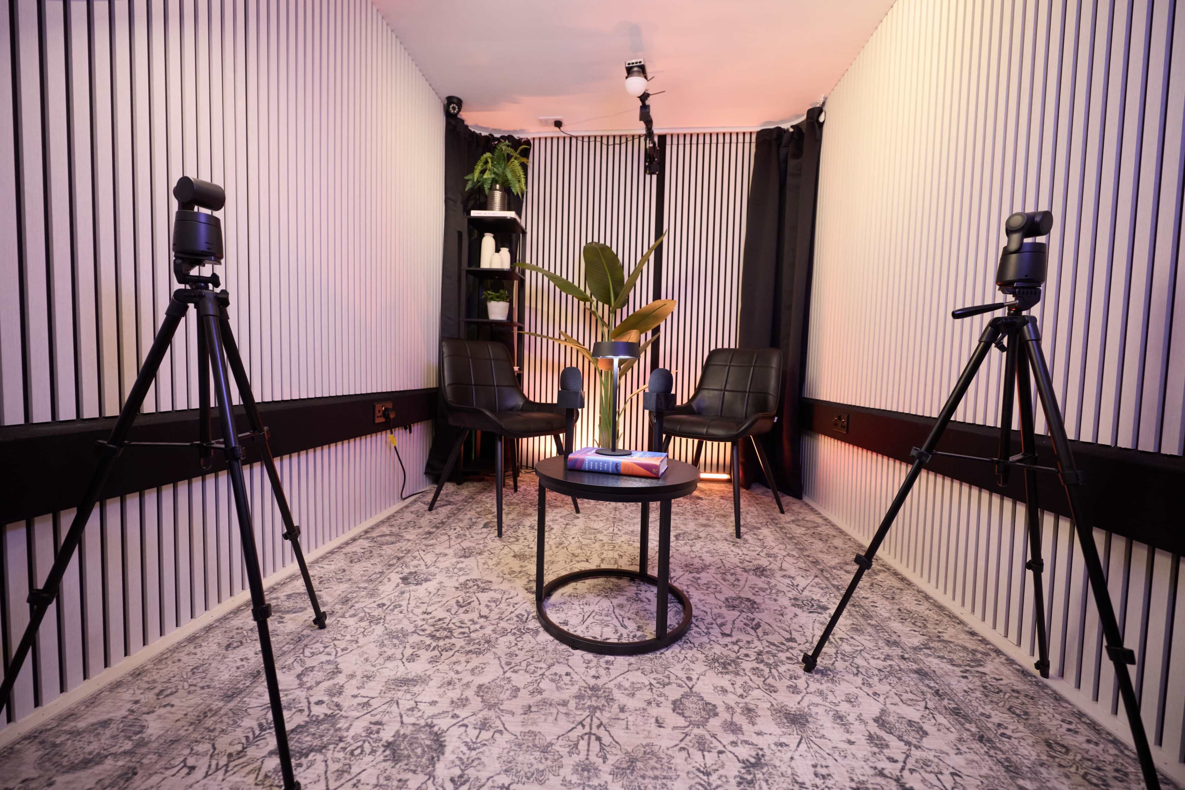 The image shows a minimalist interview setup with two chairs, a round table, and tripods for cameras, set against a striped wall and dark curtains.