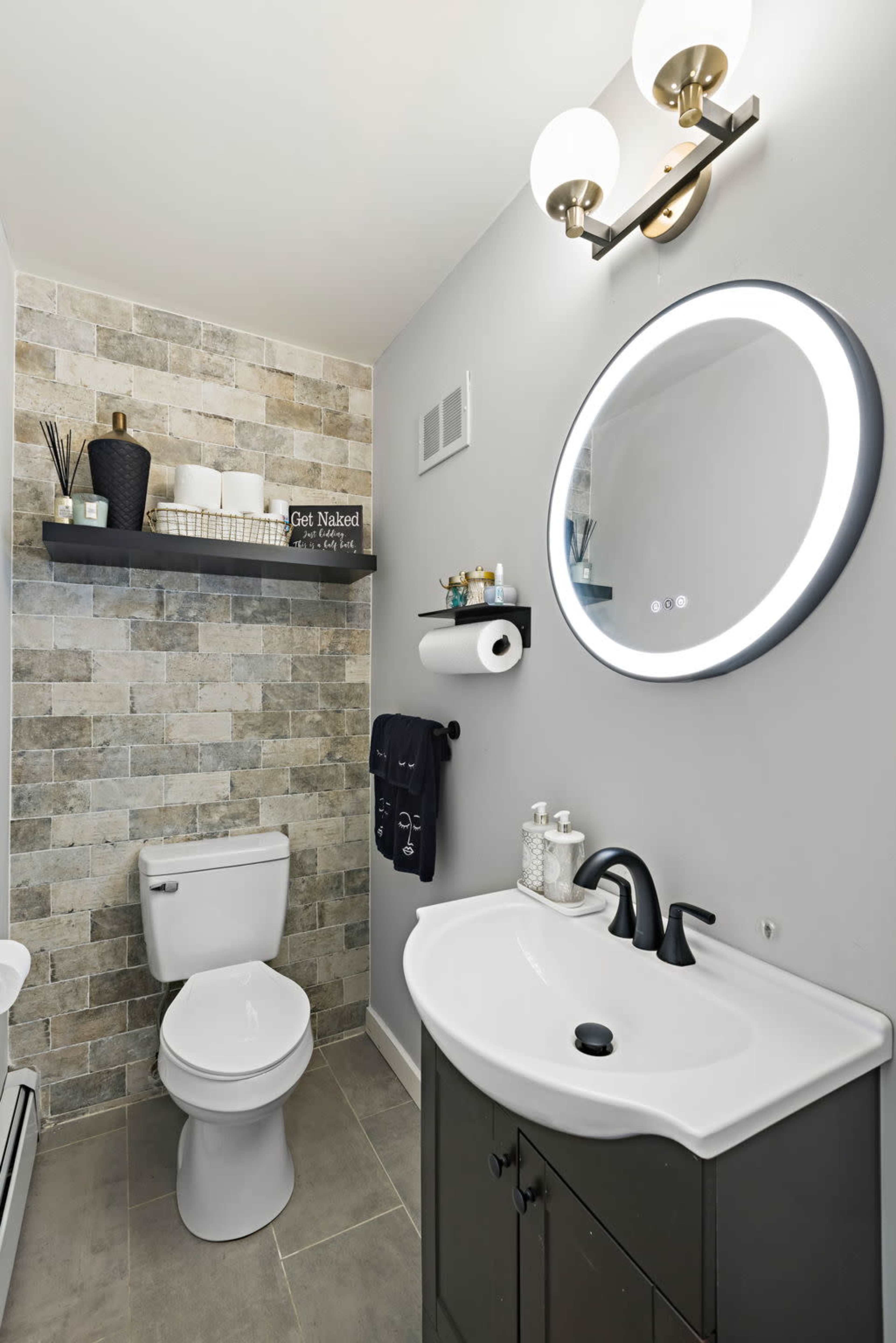 A modern bathroom featuring a wall-mounted mirror, a black vanity with a white sink, a toilet, and a shelf with decorative items and towels.