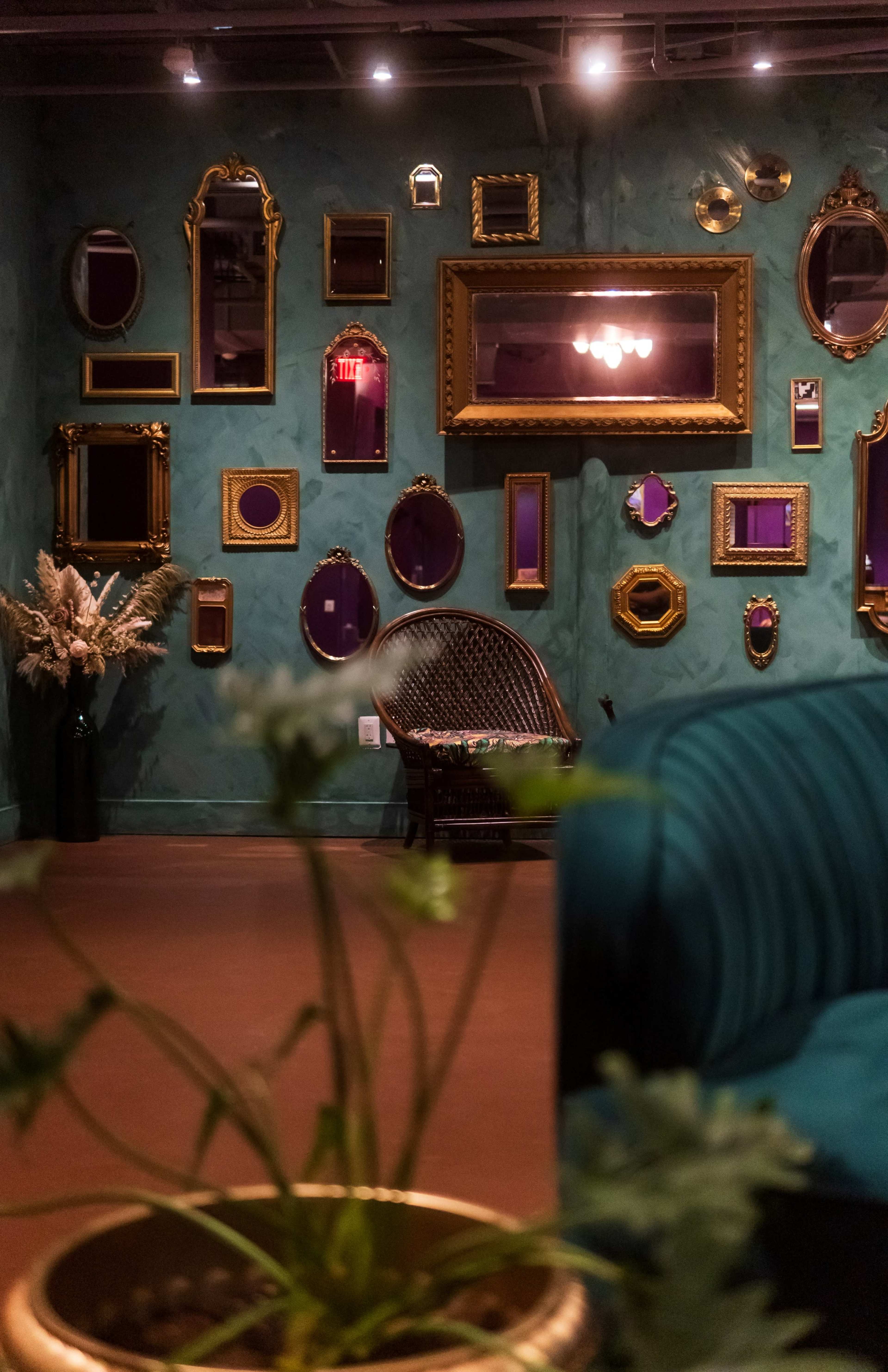 The image shows a room featuring a wall adorned with various ornate mirrors framed in gold, alongside a green couch and a small potted plant in the foreground.