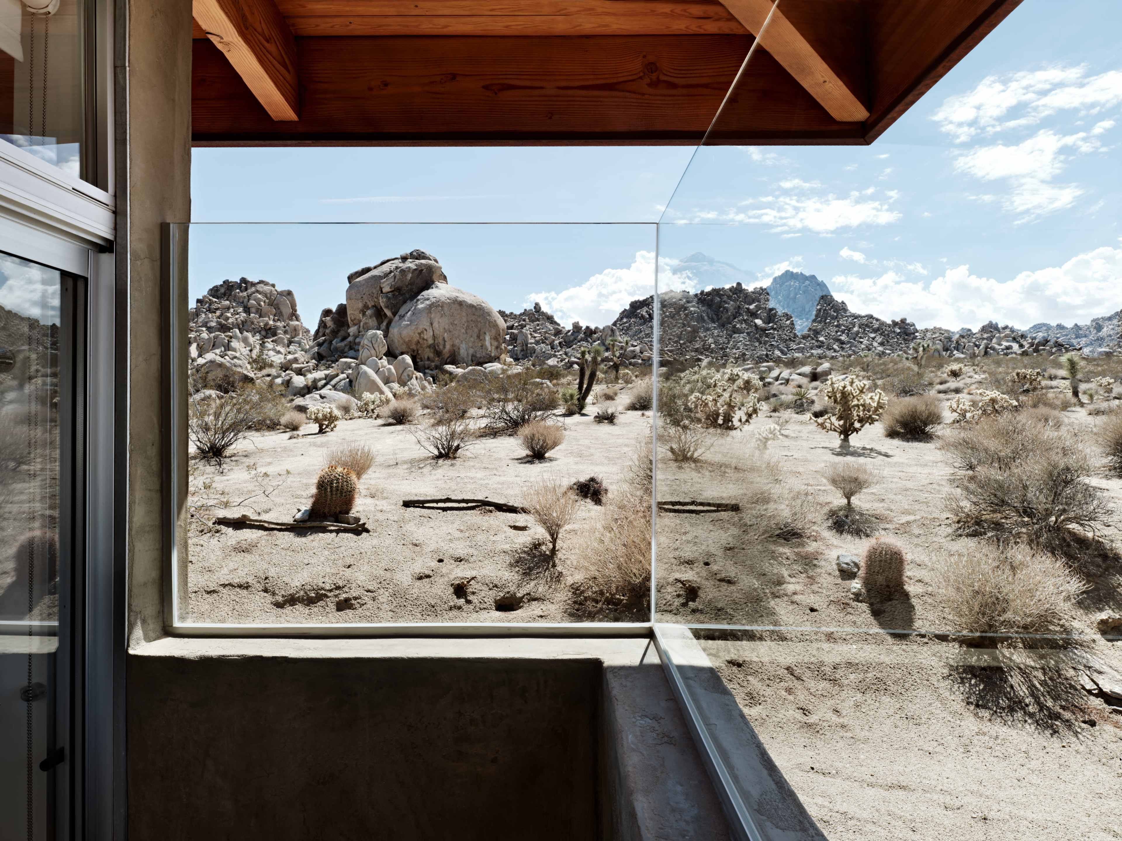 The image shows a modern architectural window framing a desert landscape with large rocks and sparse vegetation.