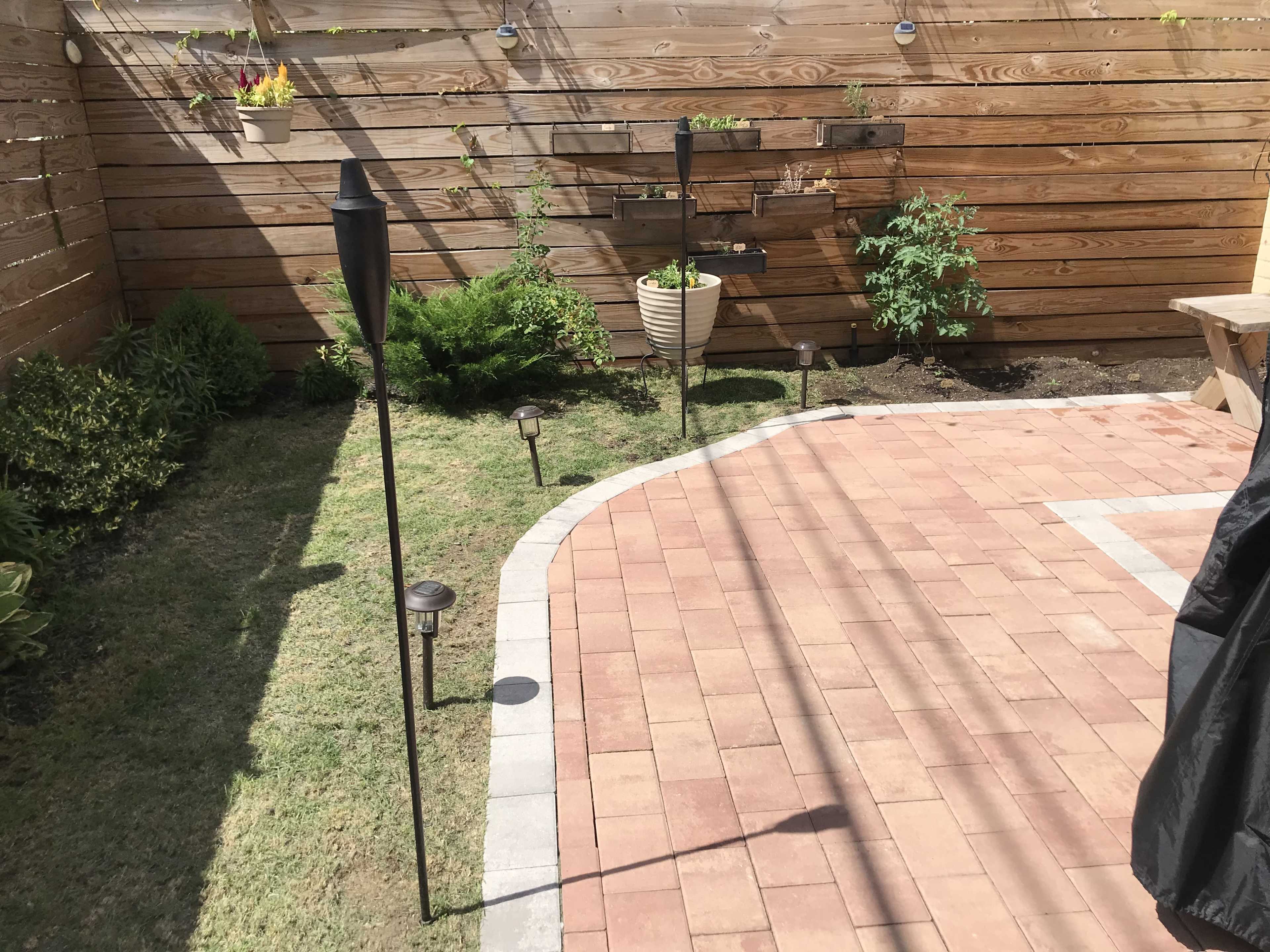The image shows a landscaped garden area with brick paving, decorative light fixtures, and wooden fencing.