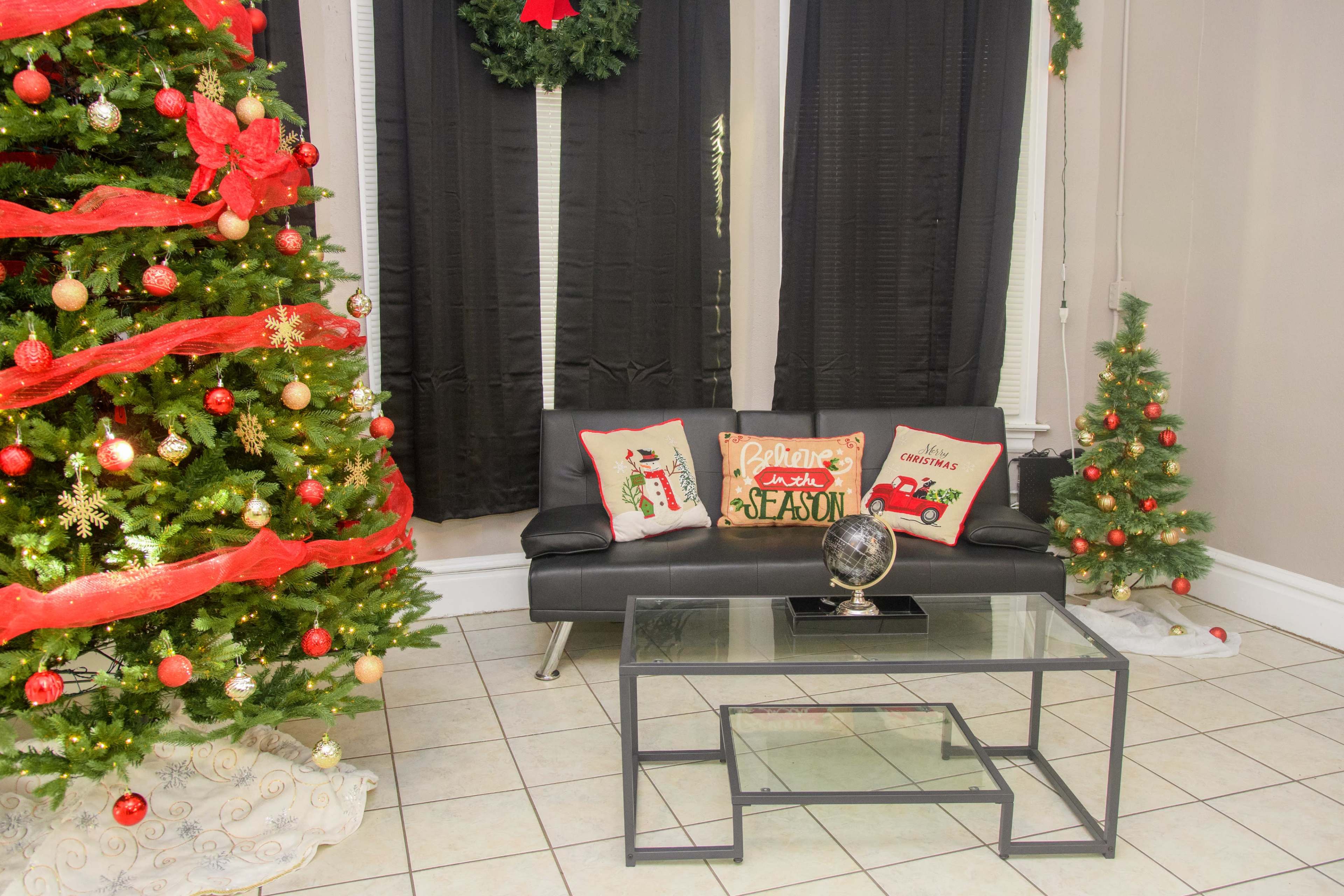 A decorated living room featuring a large Christmas tree on the left, a smaller tree on the right, and a black leather sofa with festive pillows in front of a glass coffee table.