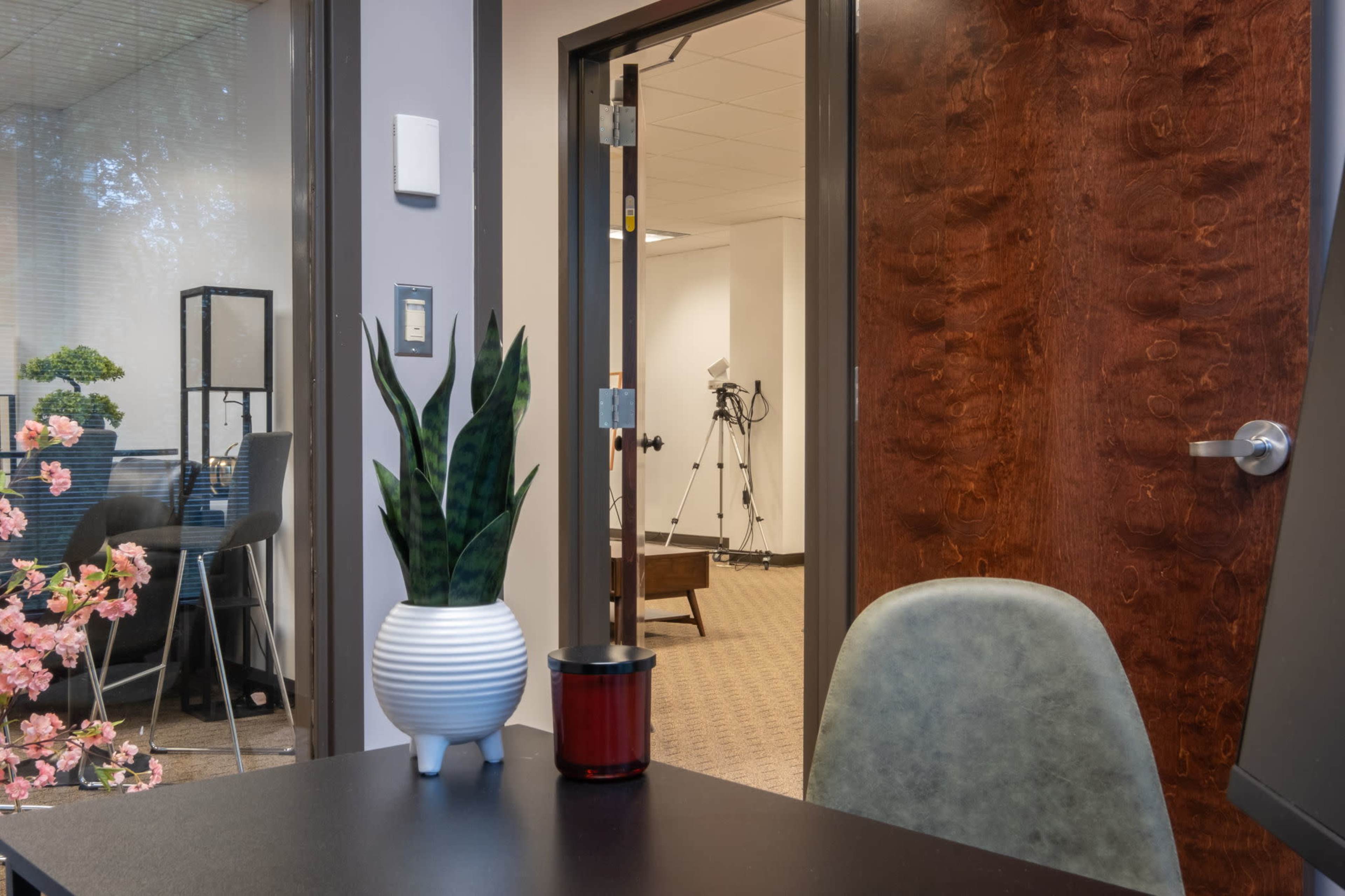 A modern office space features a desk with a plant and a candle, and a doorway leading to another room with equipment.