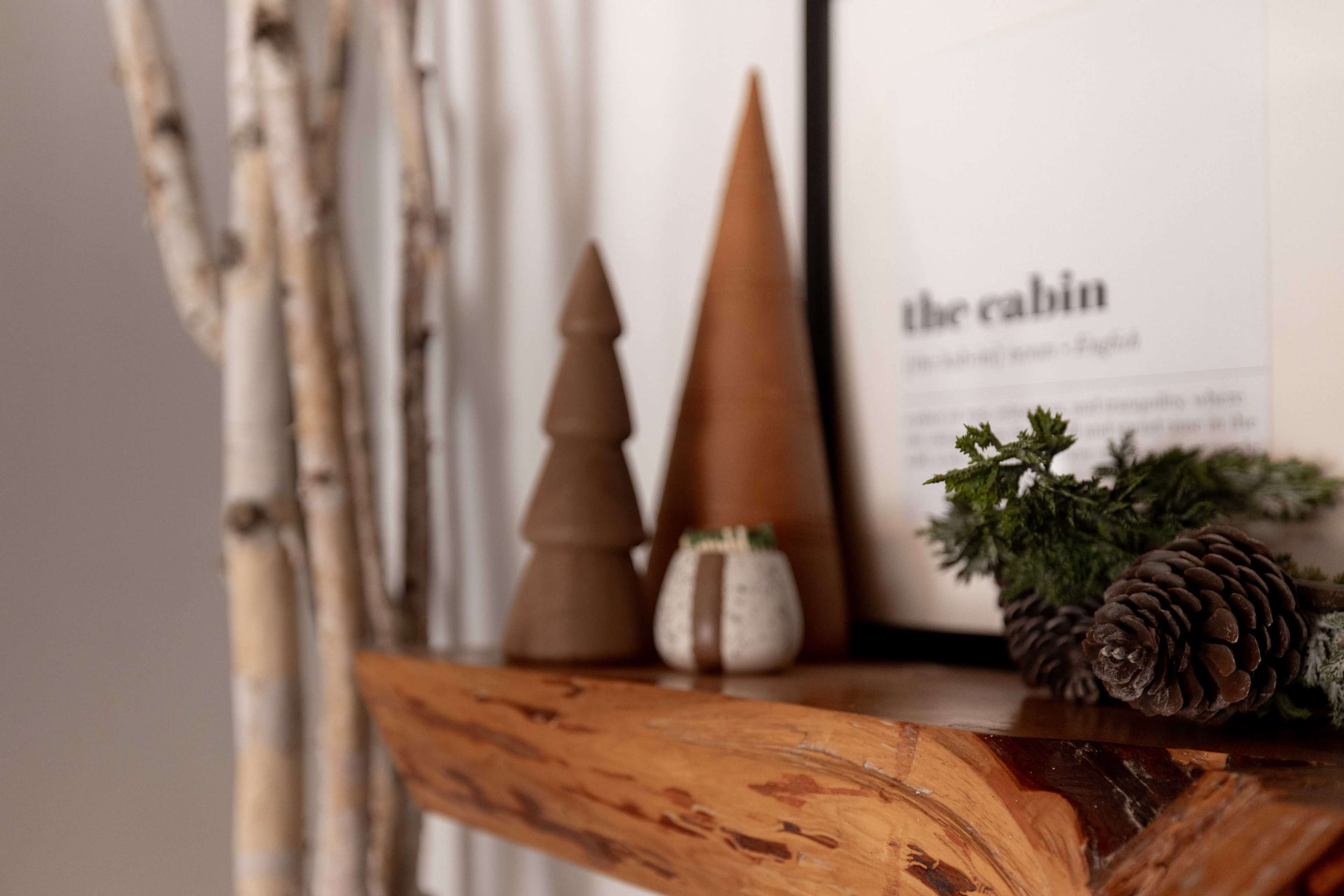 A wooden shelf displays two cone-shaped ceramic pieces, a small potted plant, pinecones, and a framed print on a wall with birch branches beside it.