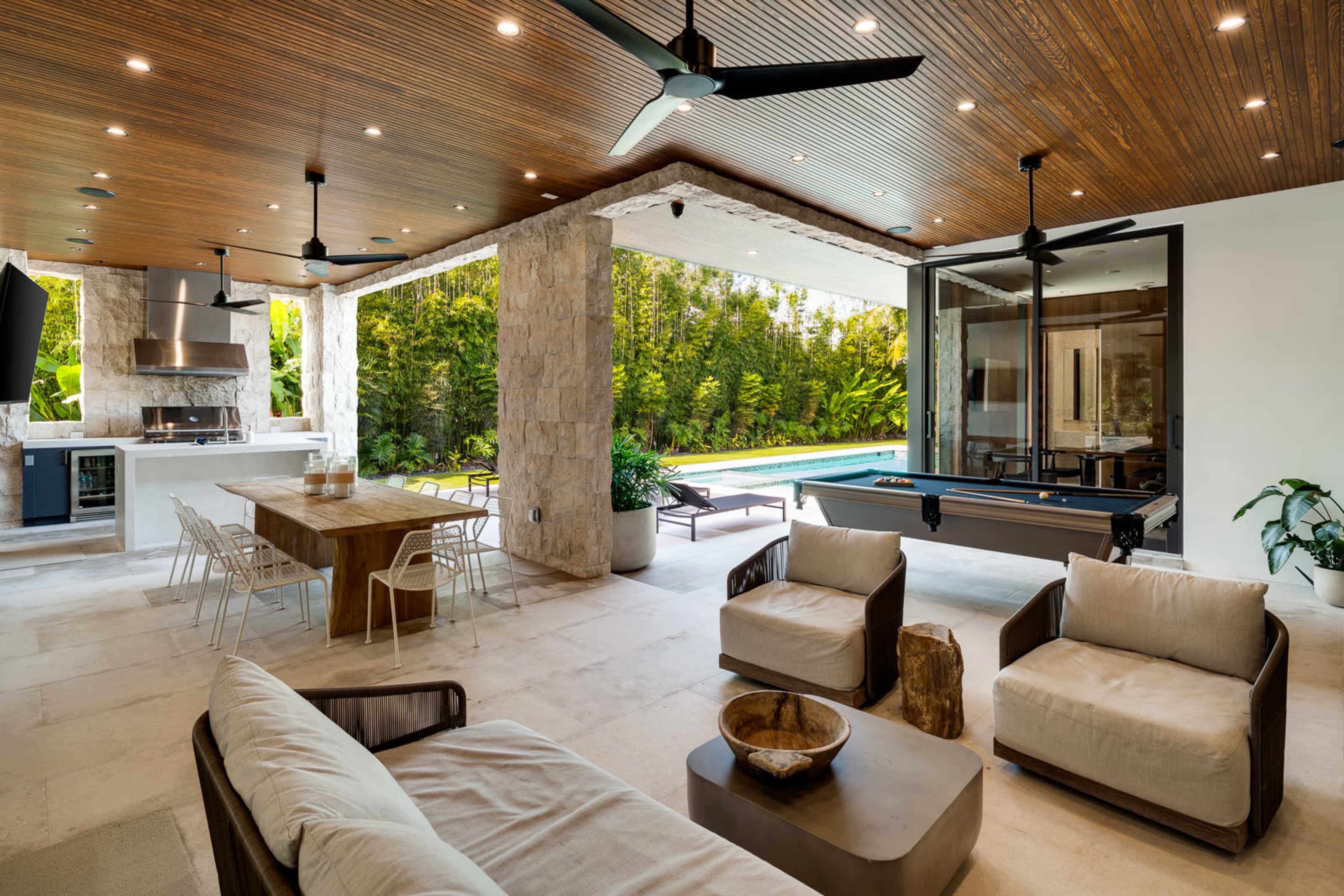 The image shows a modern outdoor patio area with a dining table, lounge seating, and a pool table, surrounded by lush greenery and featuring a swimming pool visible beyond.