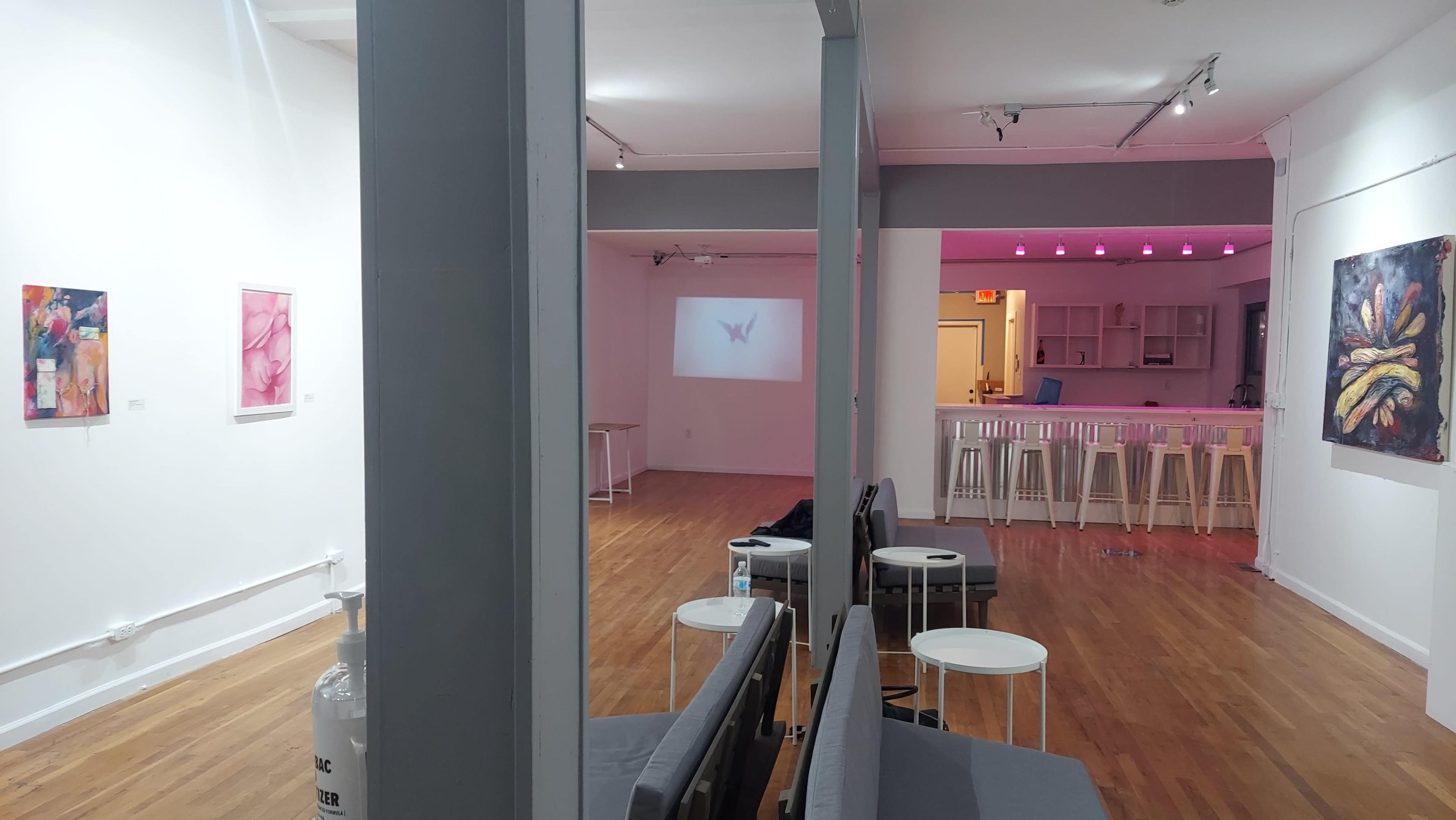 A modern gallery space with wooden floors, white walls, and a bar area in the background, featuring art pieces displayed on the walls and minimalist seating in the foreground.
