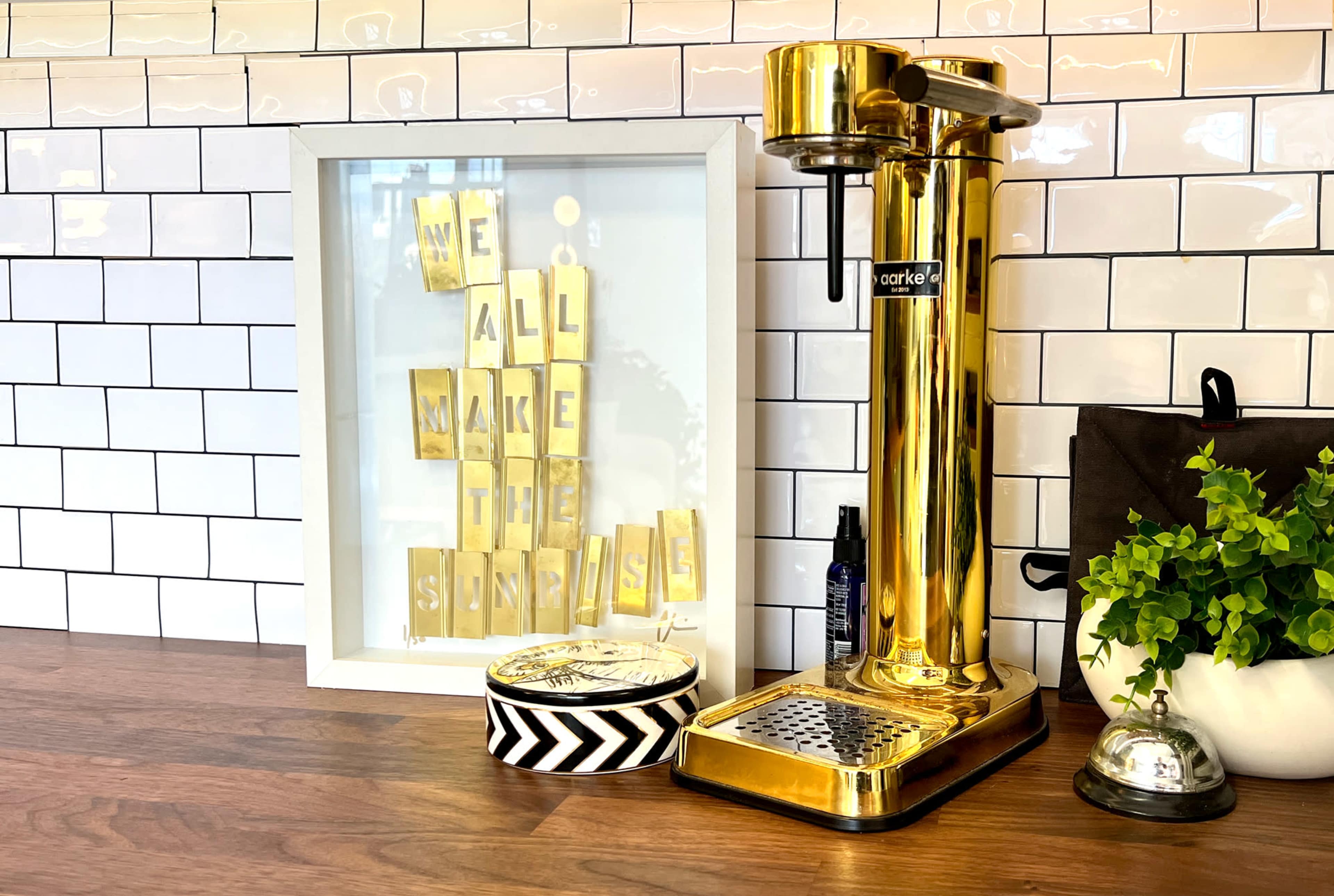 A gold-colored coffee maker stands on a wooden counter beside a framed decorative piece with the message "WE ALL MAKE THE SUNSHINE."