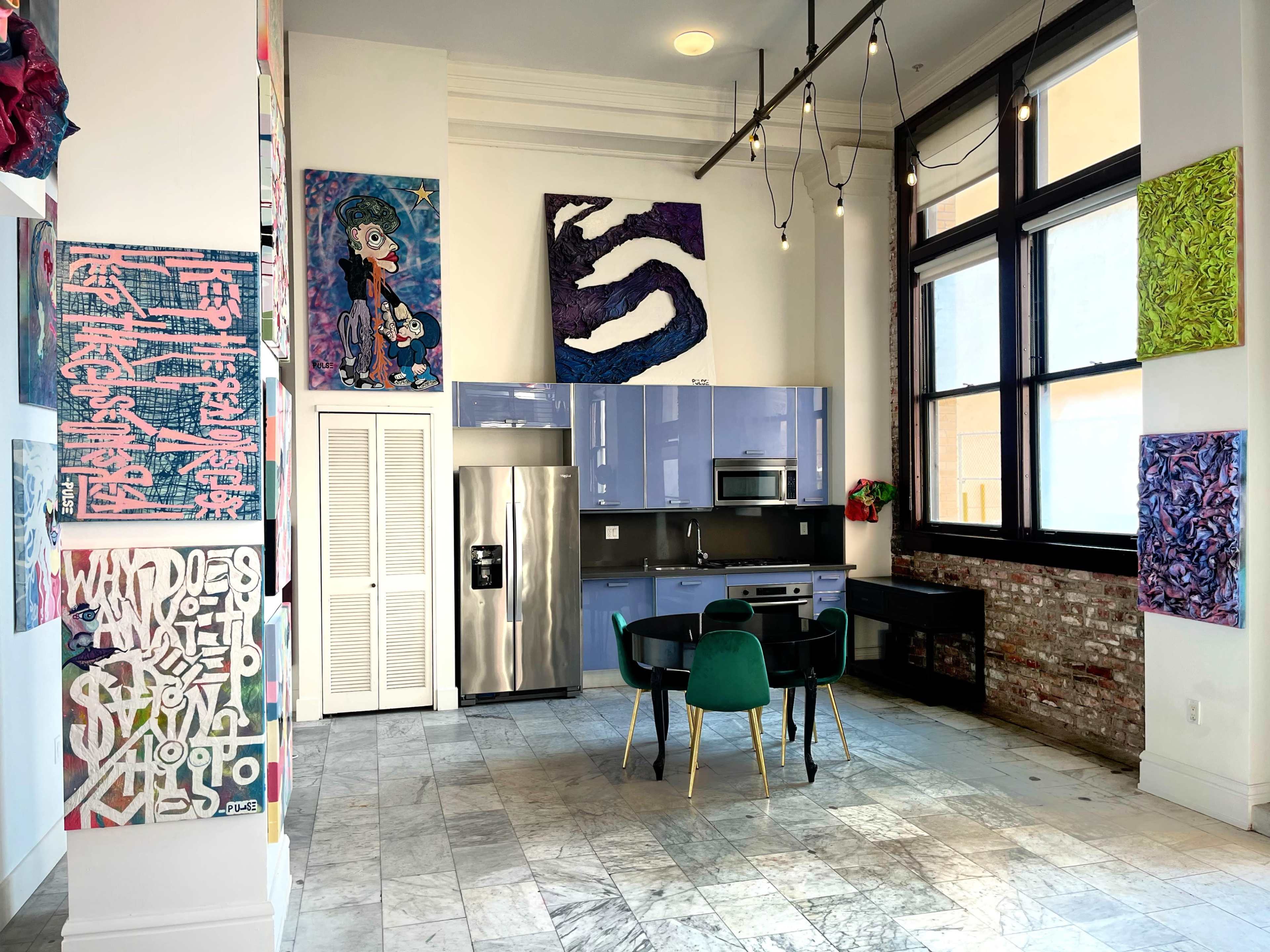 The image shows a modern kitchen space with blue cabinets, a stainless steel refrigerator, and a dining table with green chairs, surrounded by colorful artwork on the walls.