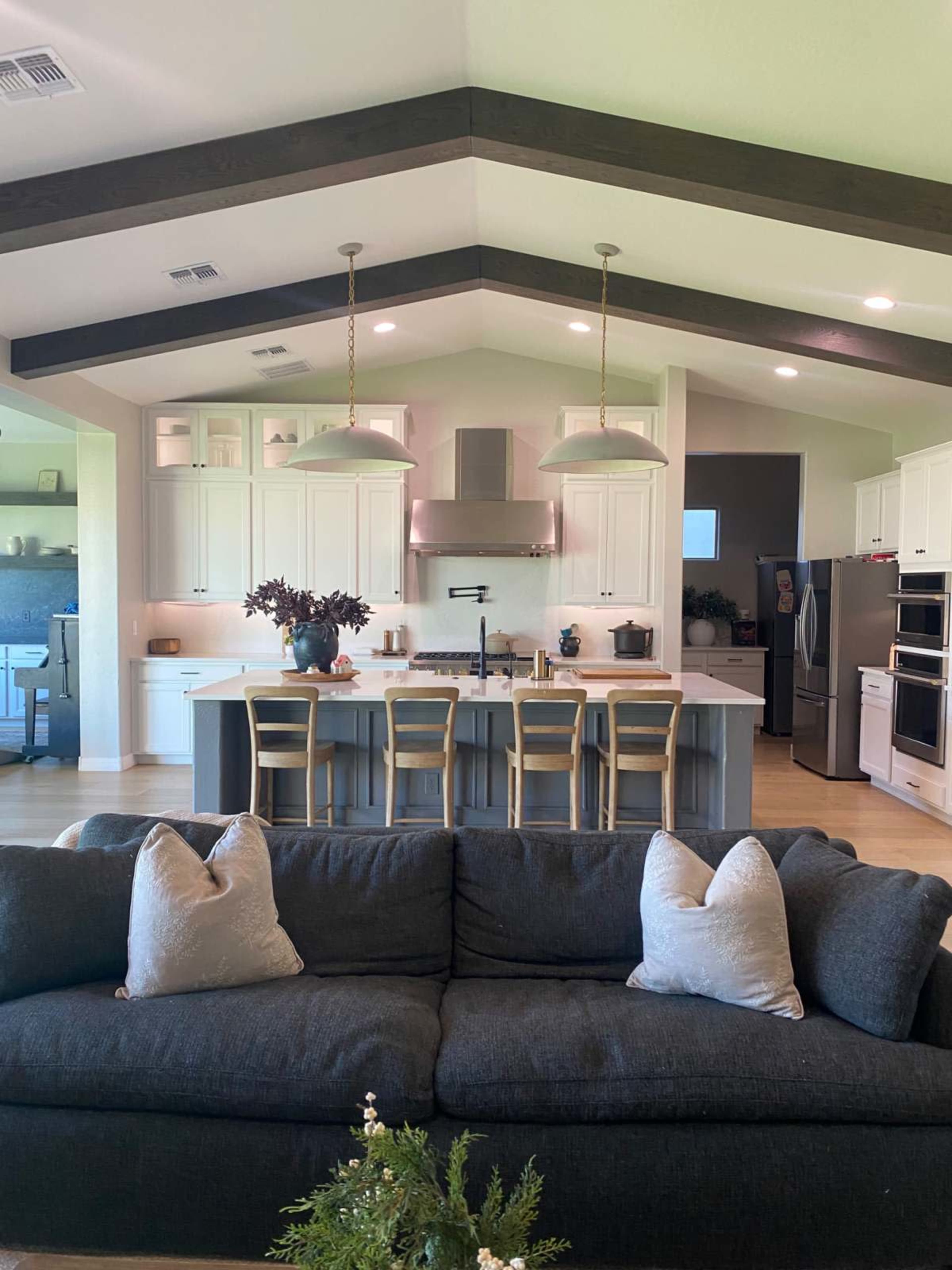 The image shows a modern kitchen with a large island, overhead pendant lights, and a cozy living area featuring a dark sofa and decorative pillows.