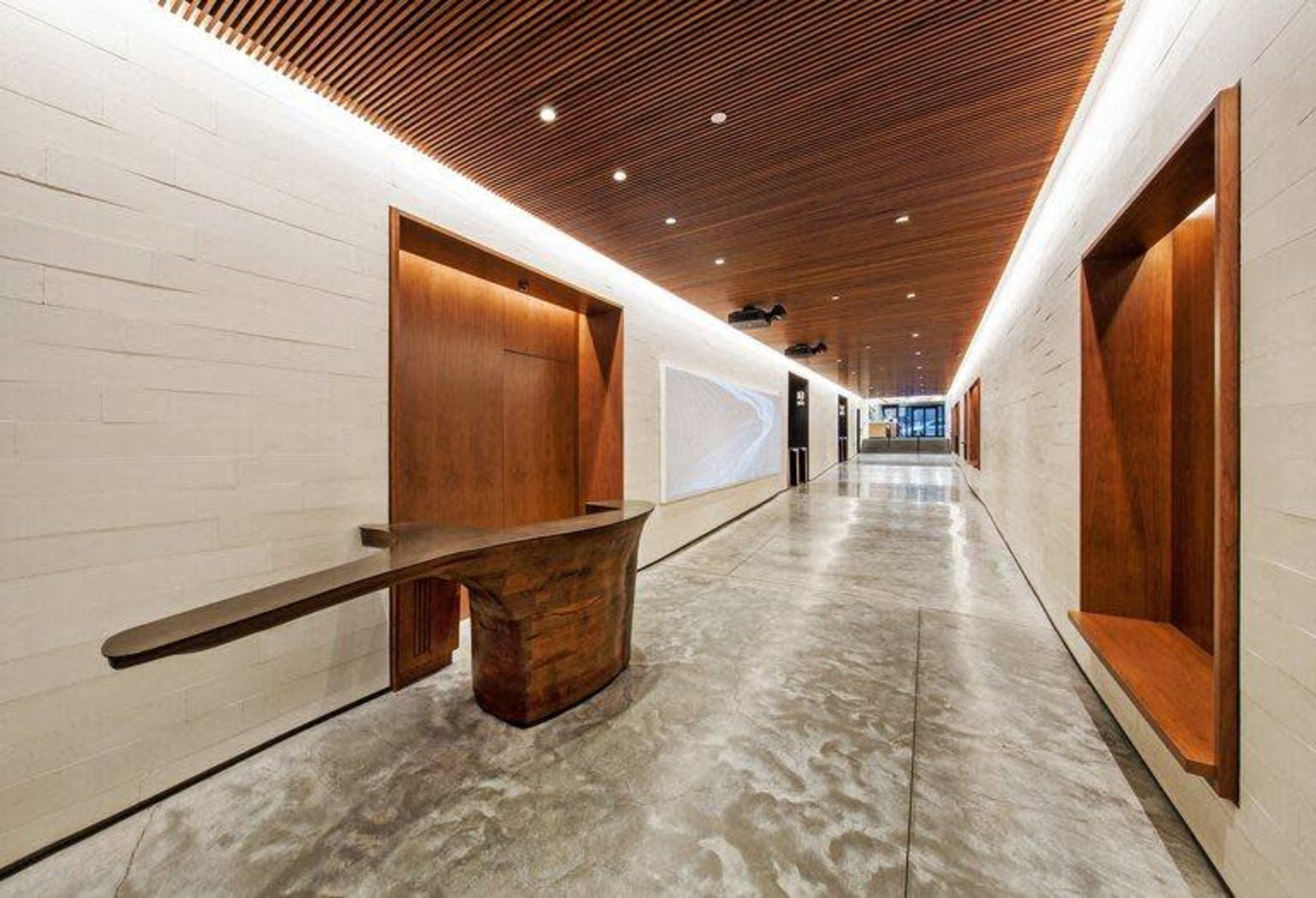 The image shows a modern hallway with smooth concrete floors, wooden paneling, and two wall recesses for display, leading to a glass entrance in the distance.
