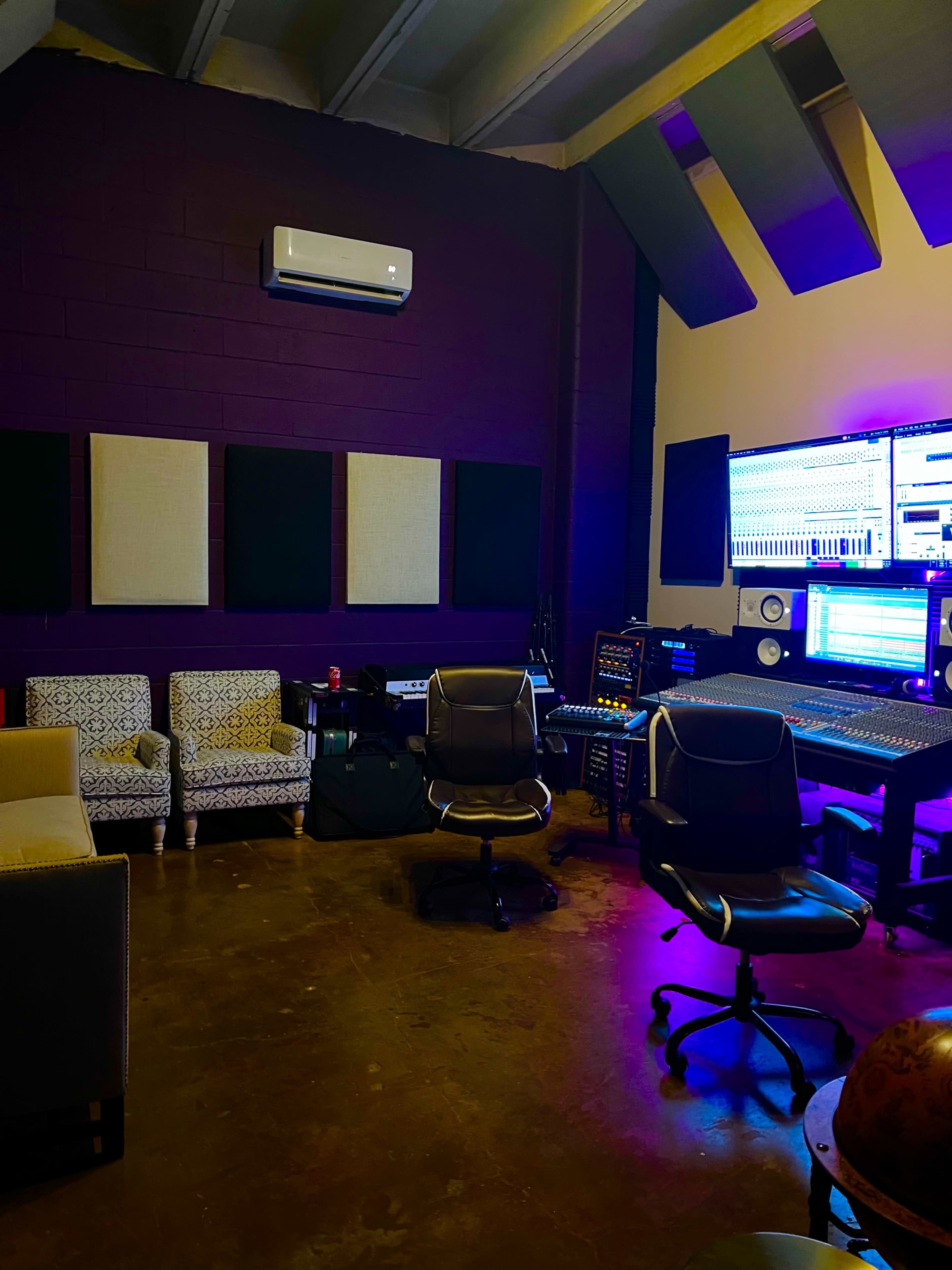 The image shows a music production studio featuring a large mixing console, multiple screens, and seating areas with two black office chairs and a beige sofa.
