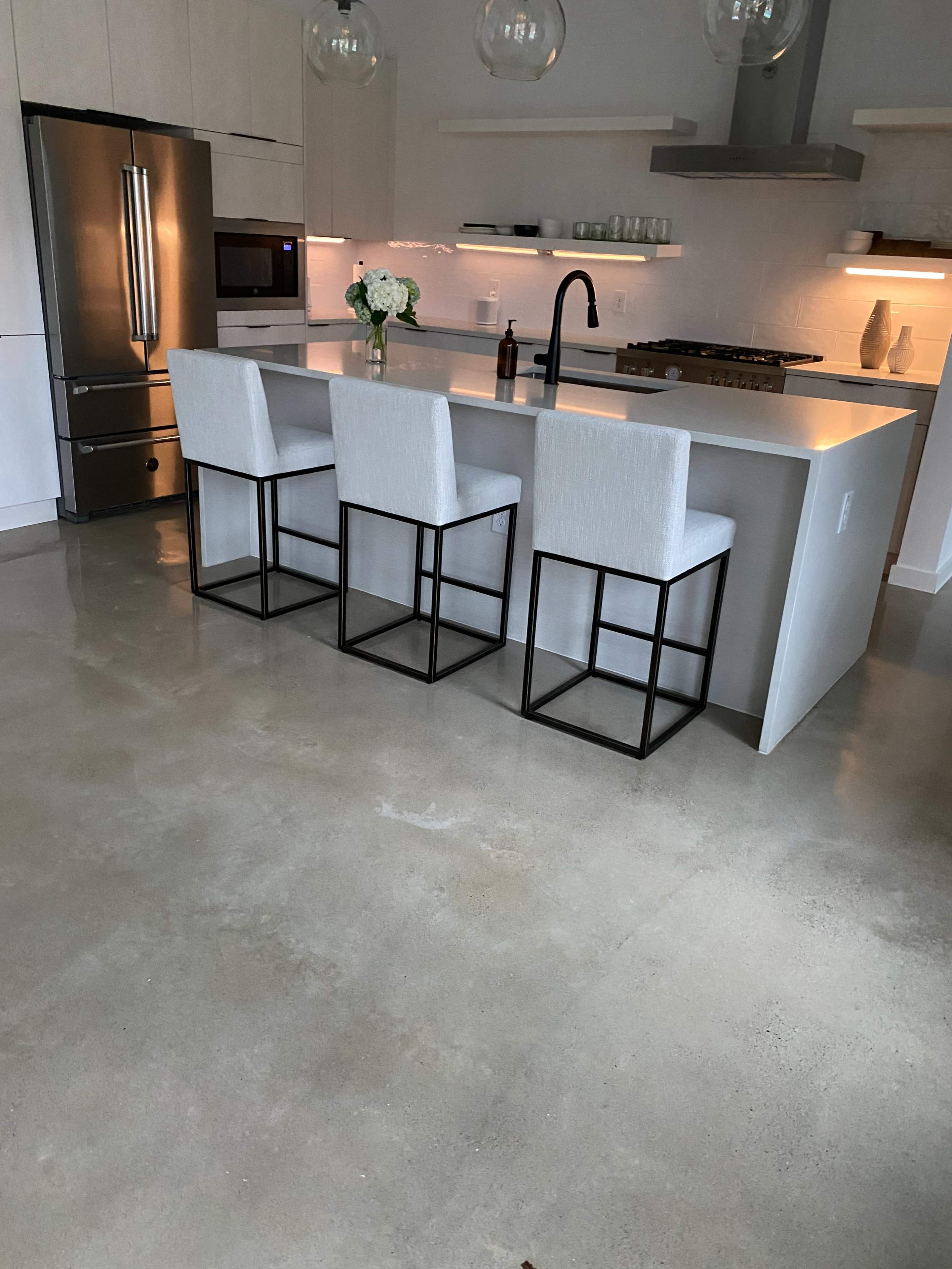 The image shows a modern kitchen featuring a large island with three bar stools, a stainless steel refrigerator, and soft ambient lighting.