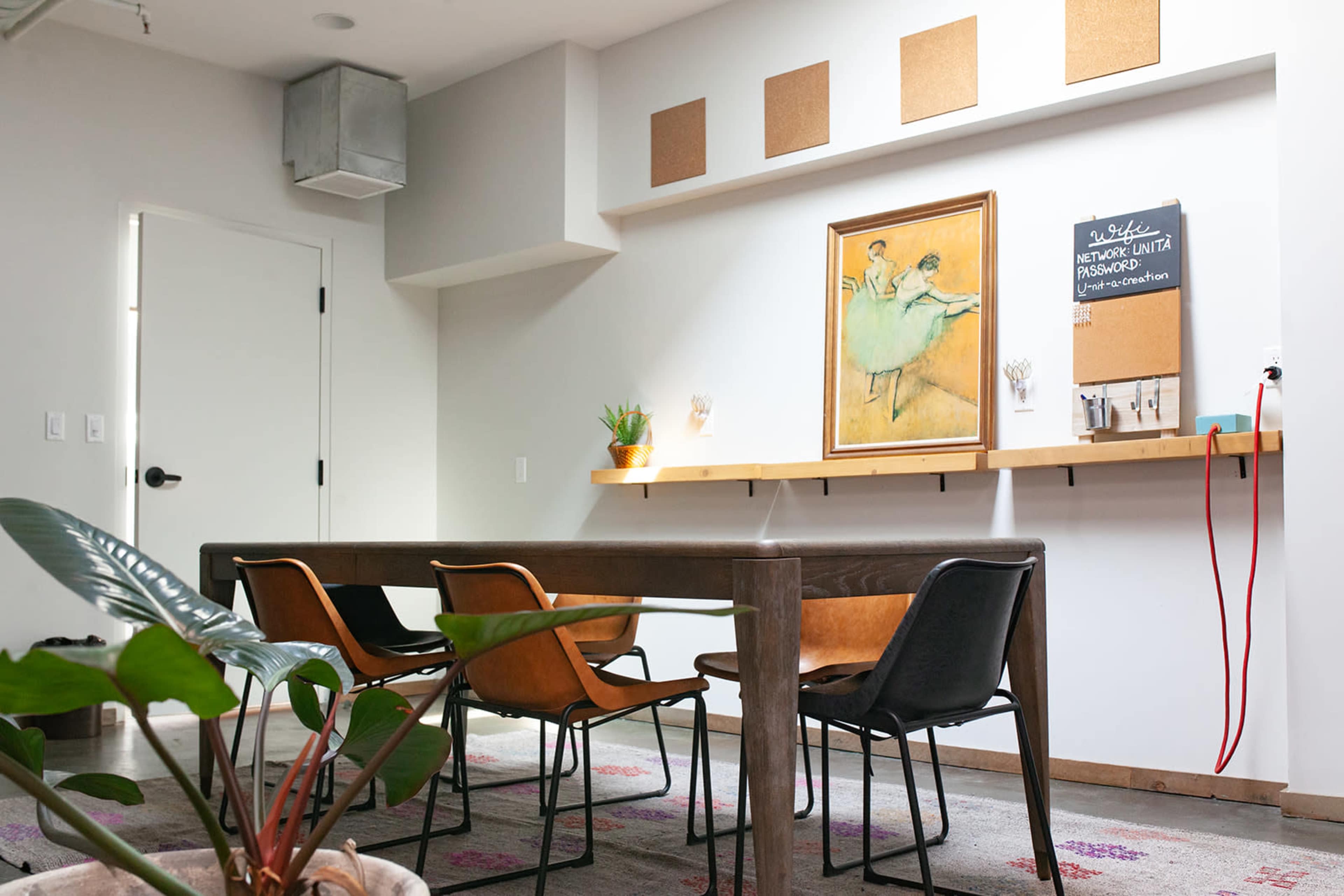 A modern workspace features a long wooden table, four chairs, a corkboard wall, a painting of two dancers, and a potted plant.
