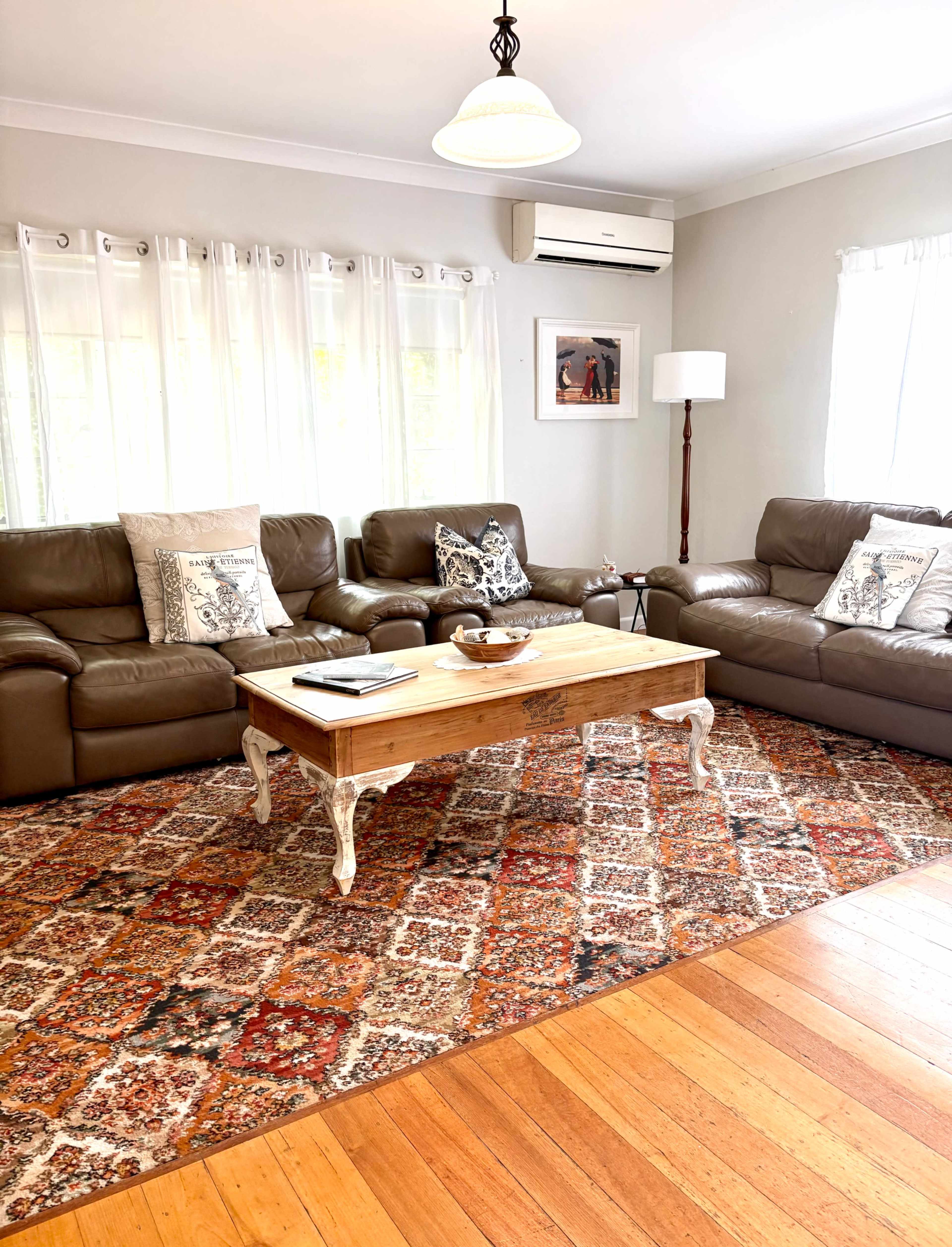 The living room features brown leather sofas arranged around a wooden coffee table on an ornate area rug, with large windows and light curtains allowing natural light to flood the space.