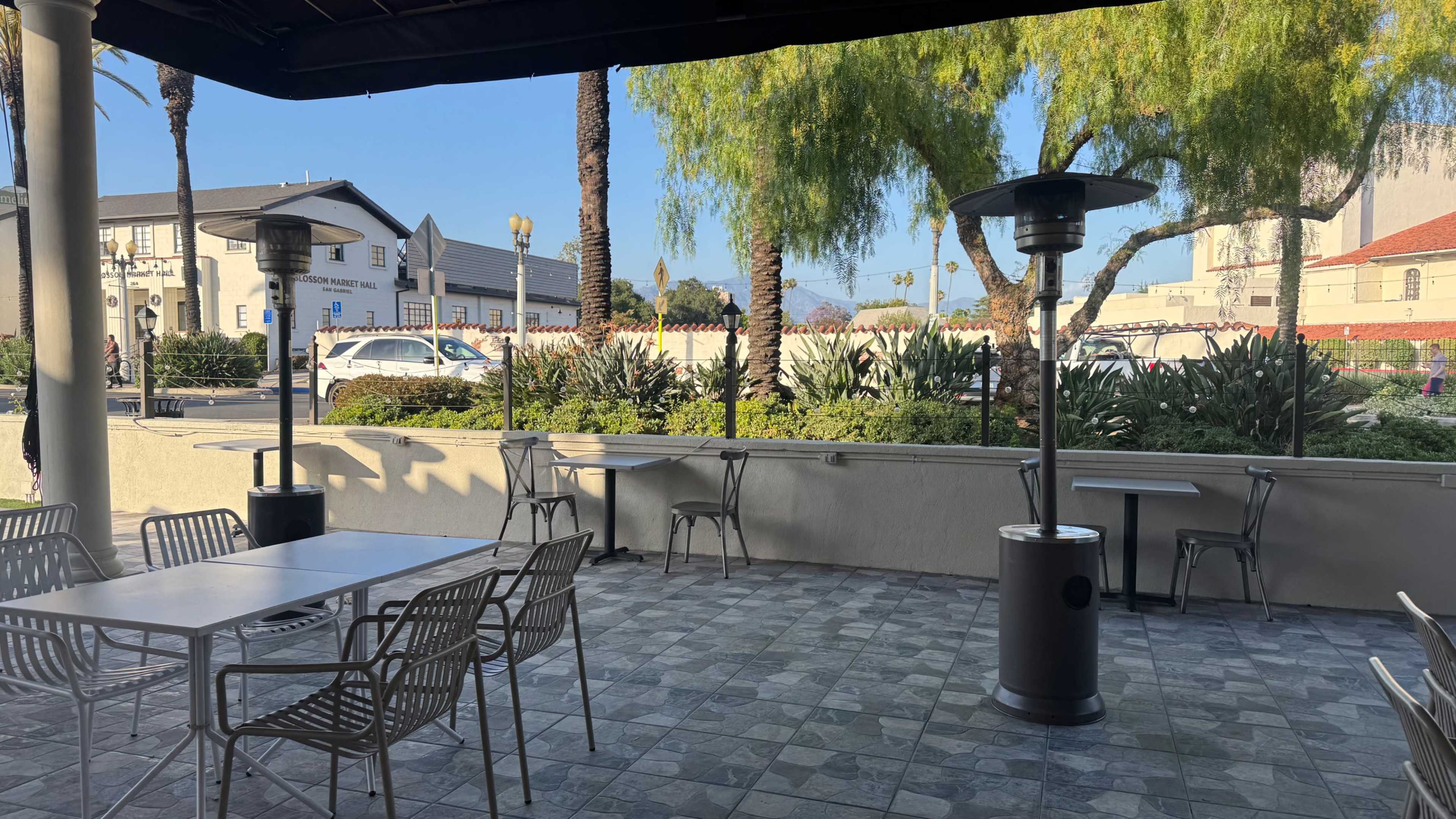 The image shows an outdoor seating area with metal tables and chairs, two patio heaters, and a landscaped view featuring palm trees and greenery.