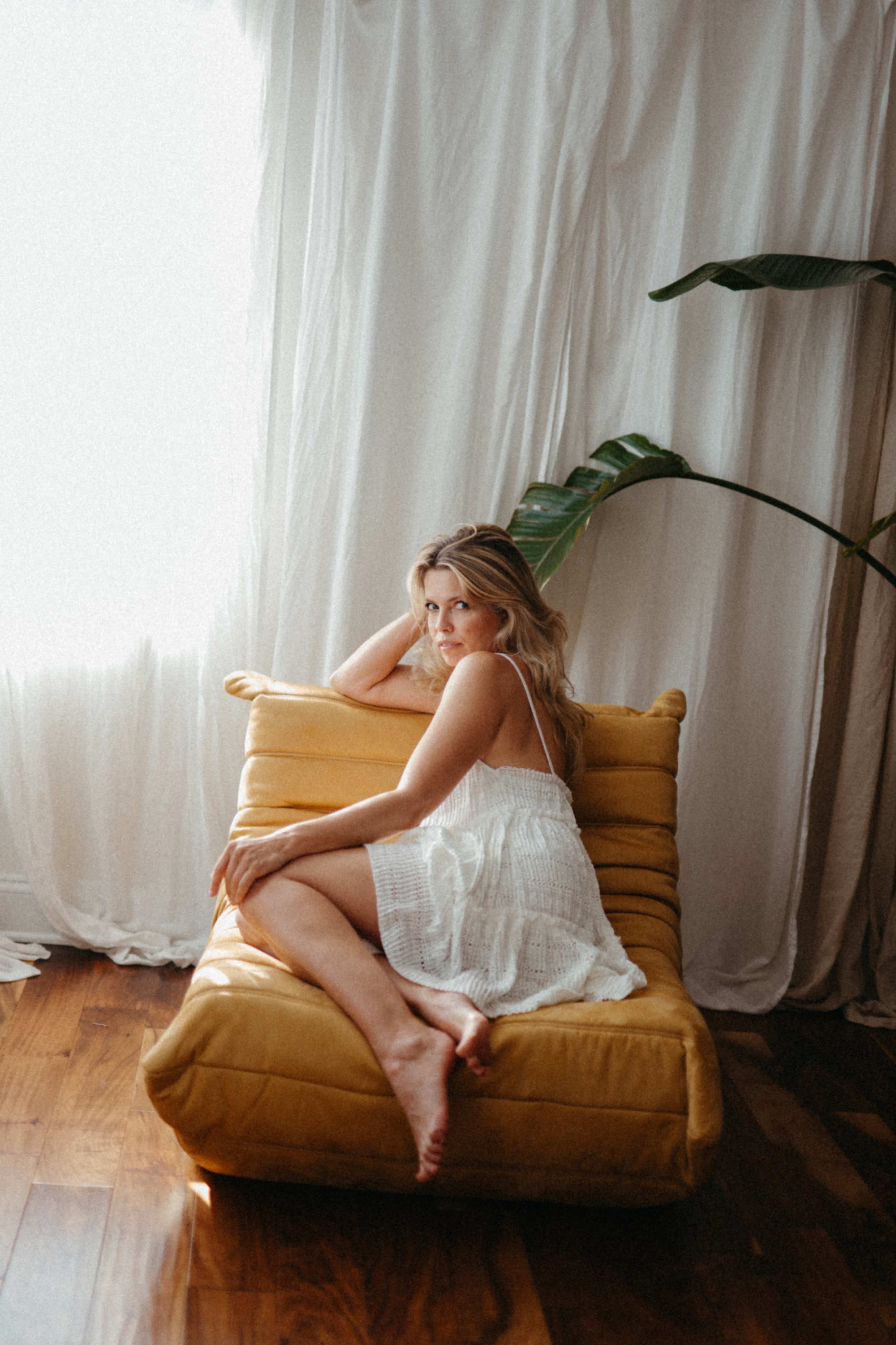 A woman in a white dress reclines on a yellow lounge chair near a window with sheer curtains.