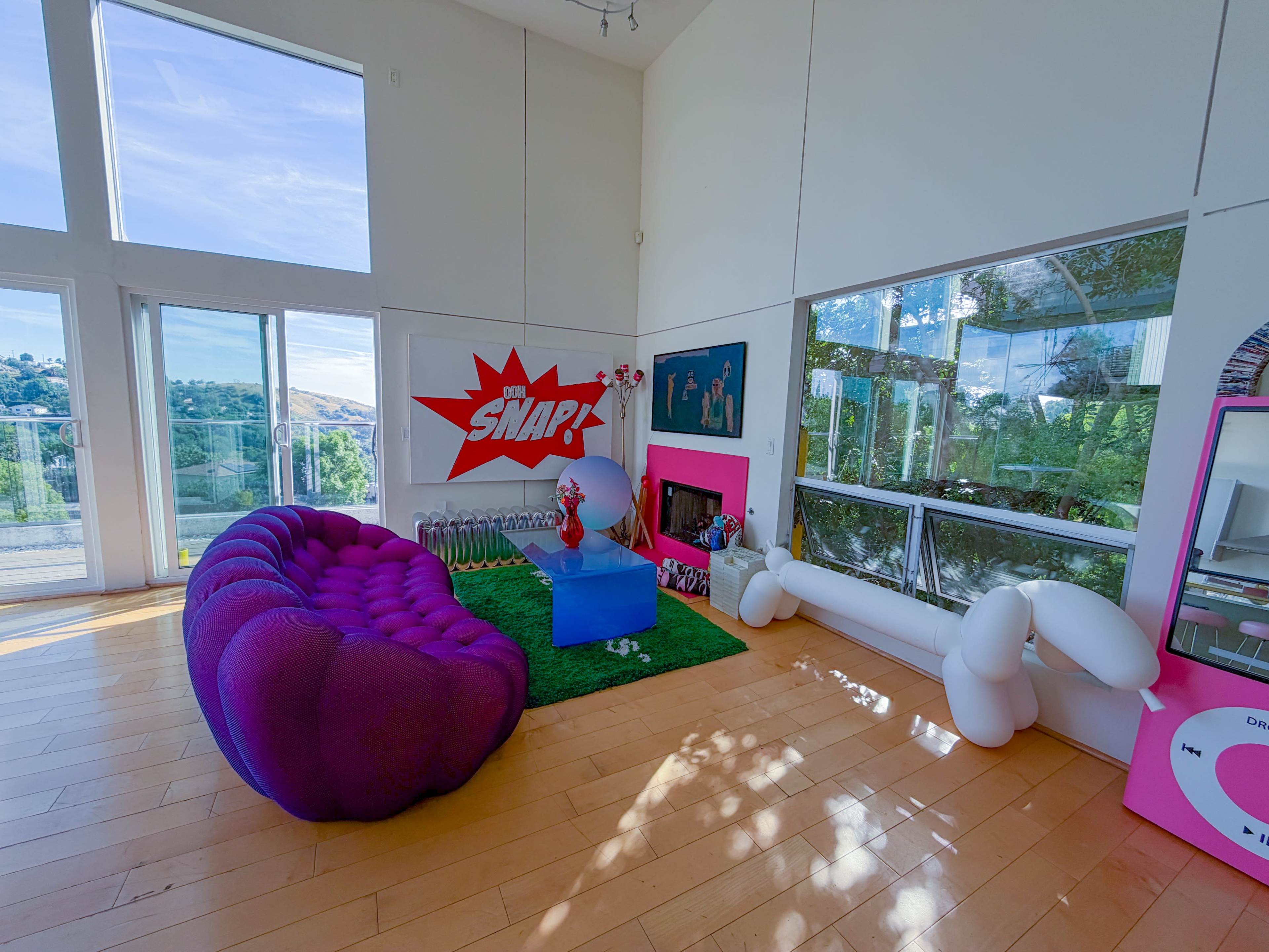 The image shows a modern living room with large windows, featuring a purple tufted sofa, green decorative grass, and colorful wall artwork.