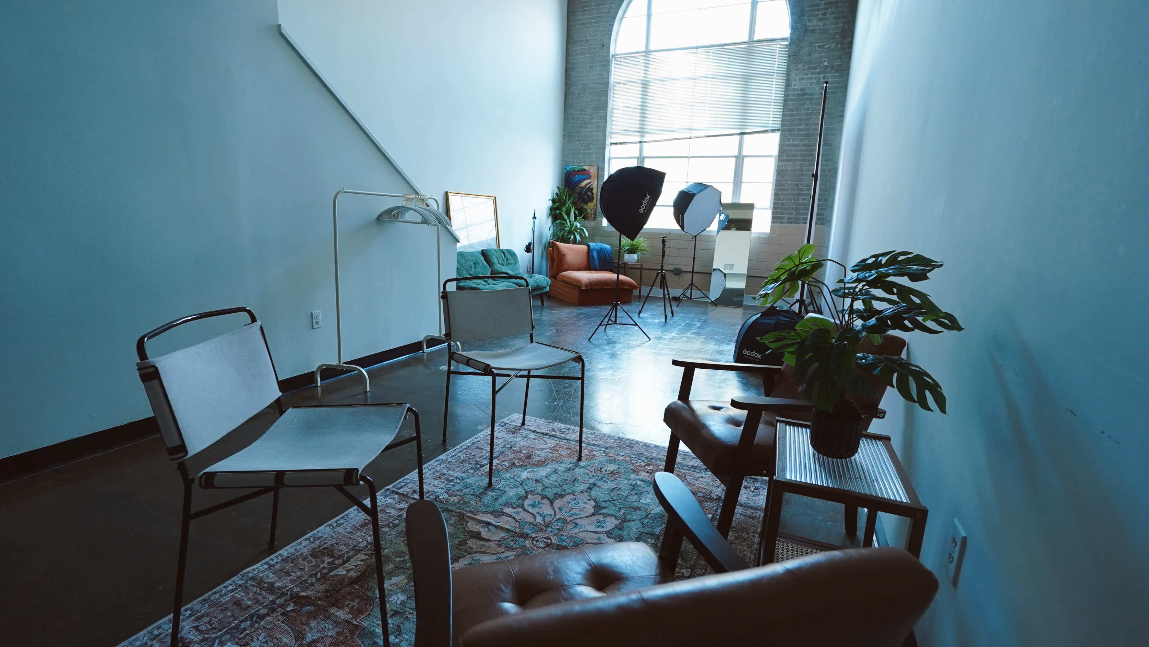 The image shows a spacious room with a mix of seating options, including chairs and a couch, alongside photography equipment and plants.