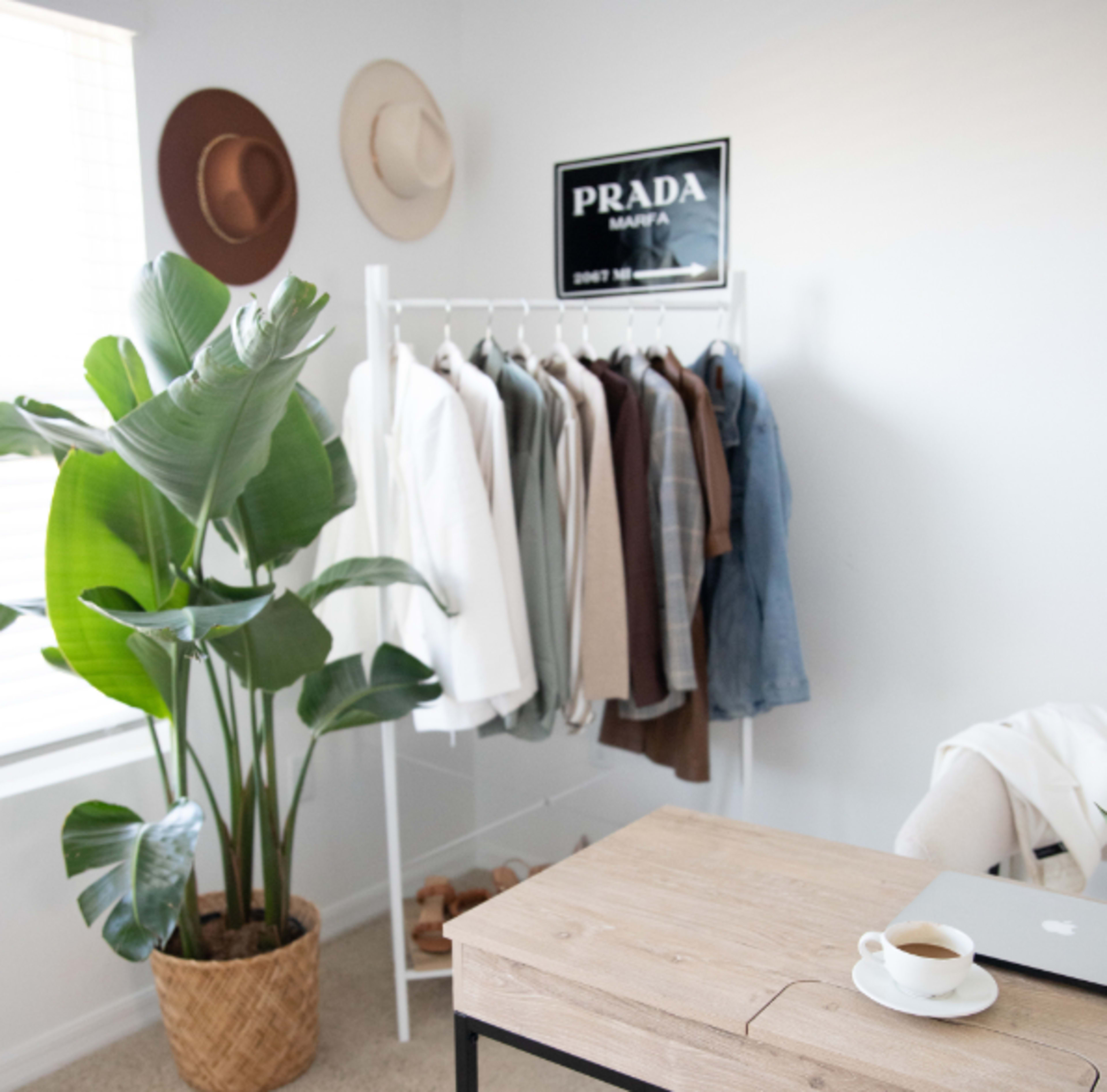A minimalist office space features a clothing rack with various garments, a potted plant, a wooden desk, and a mug on the desk.