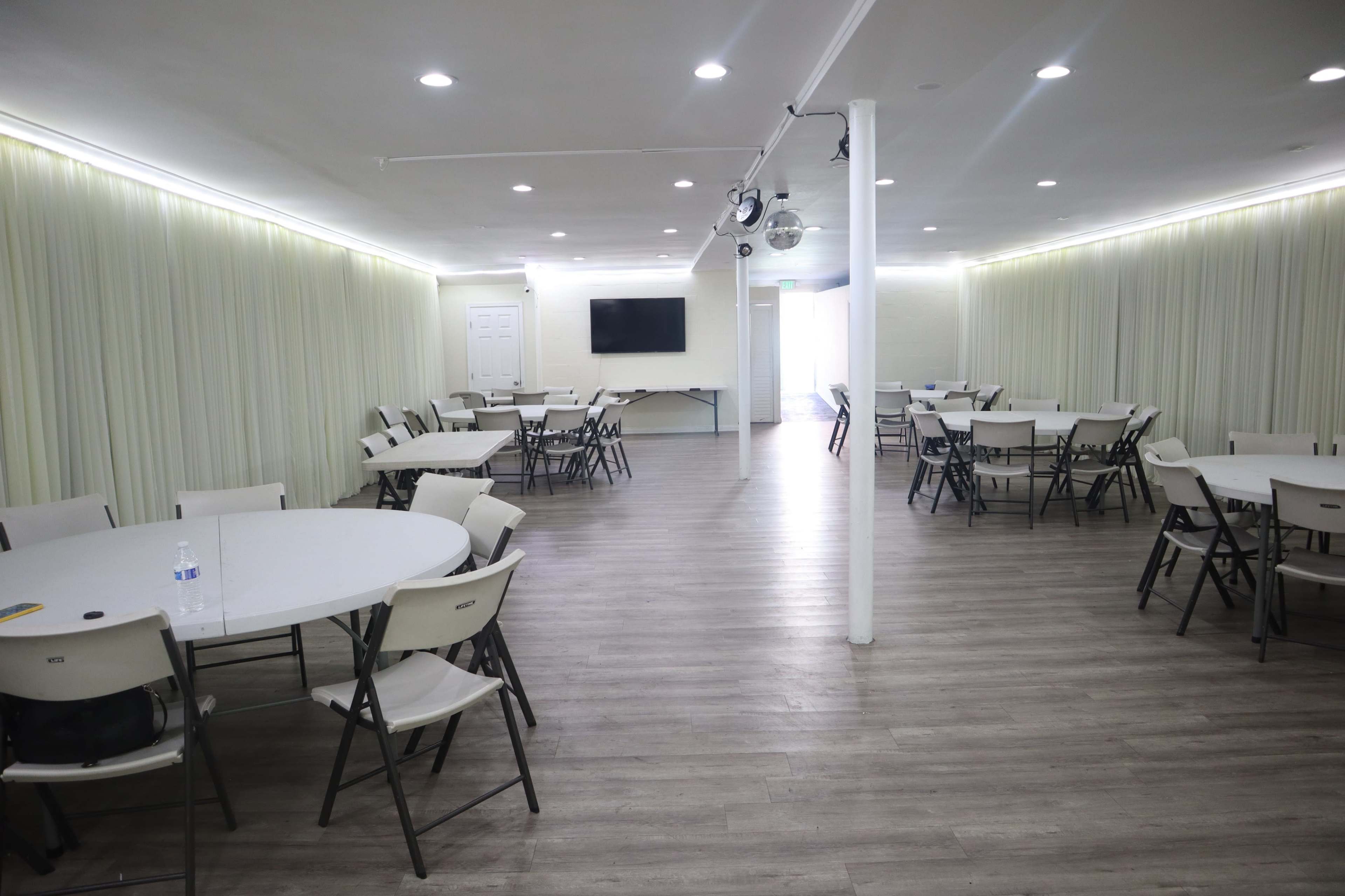 The room features multiple circular tables surrounded by chairs, with a large screen on one wall and soft lighting from the curtain-covered walls.