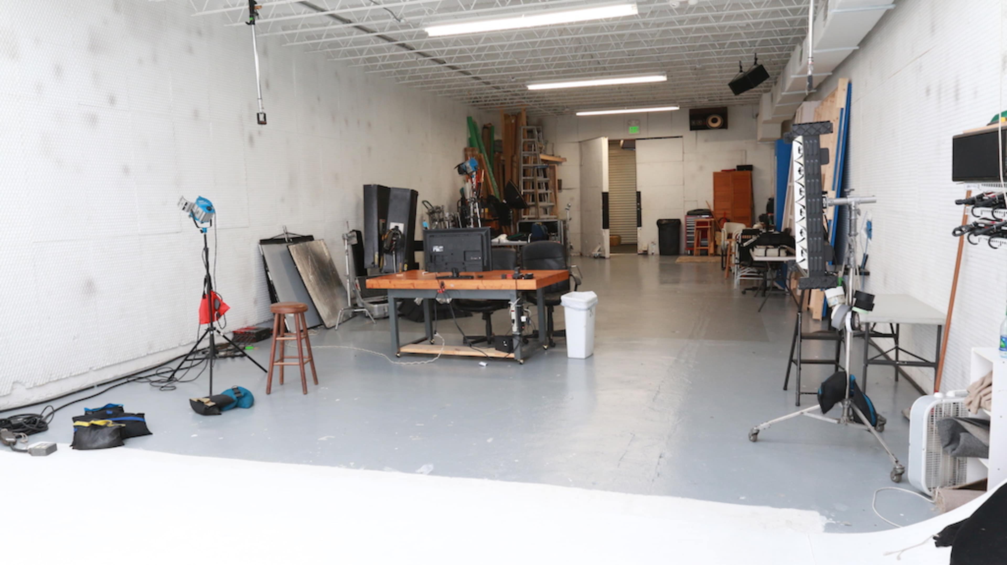 The image shows a spacious, well-lit studio with a work desk, various lighting equipment, and storage shelves along the walls.