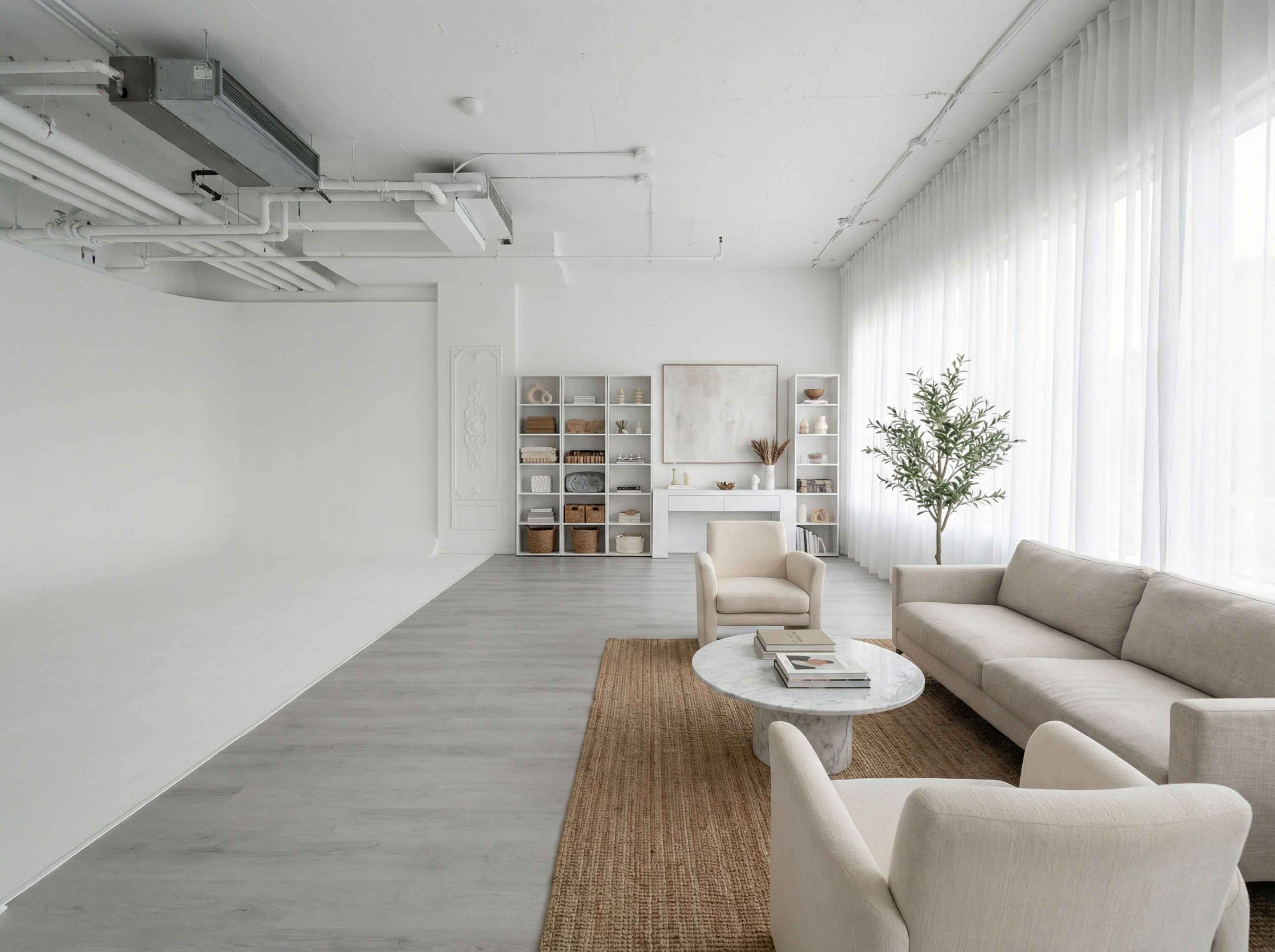 The image shows a spacious, modern living area with light-colored furniture, a round coffee table, and shelves filled with decorative items against a backdrop of large windows.