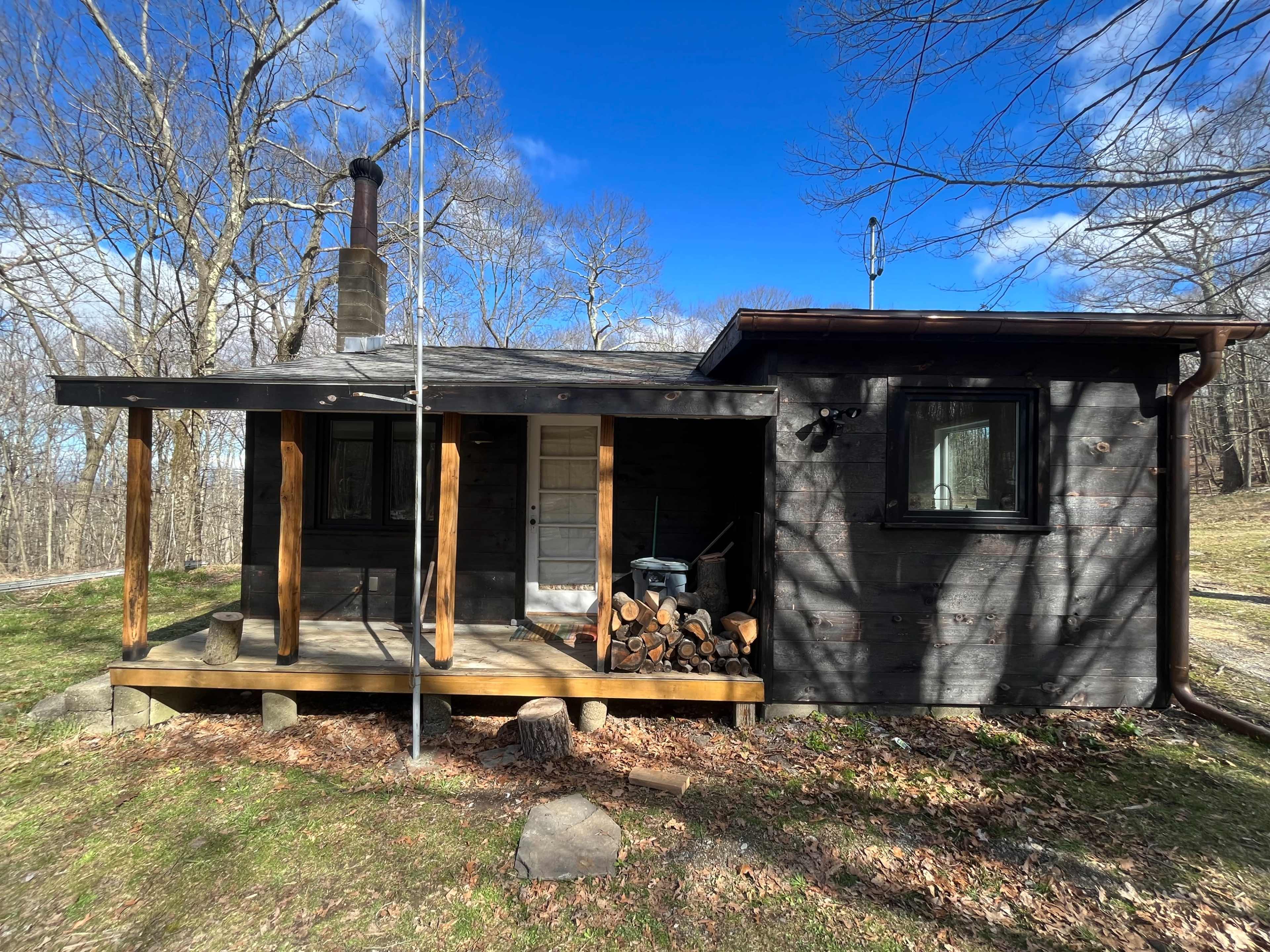 A wooden cabin with a porch, stacked firewood, and a chimney, set in a forested area under a partly cloudy sky.