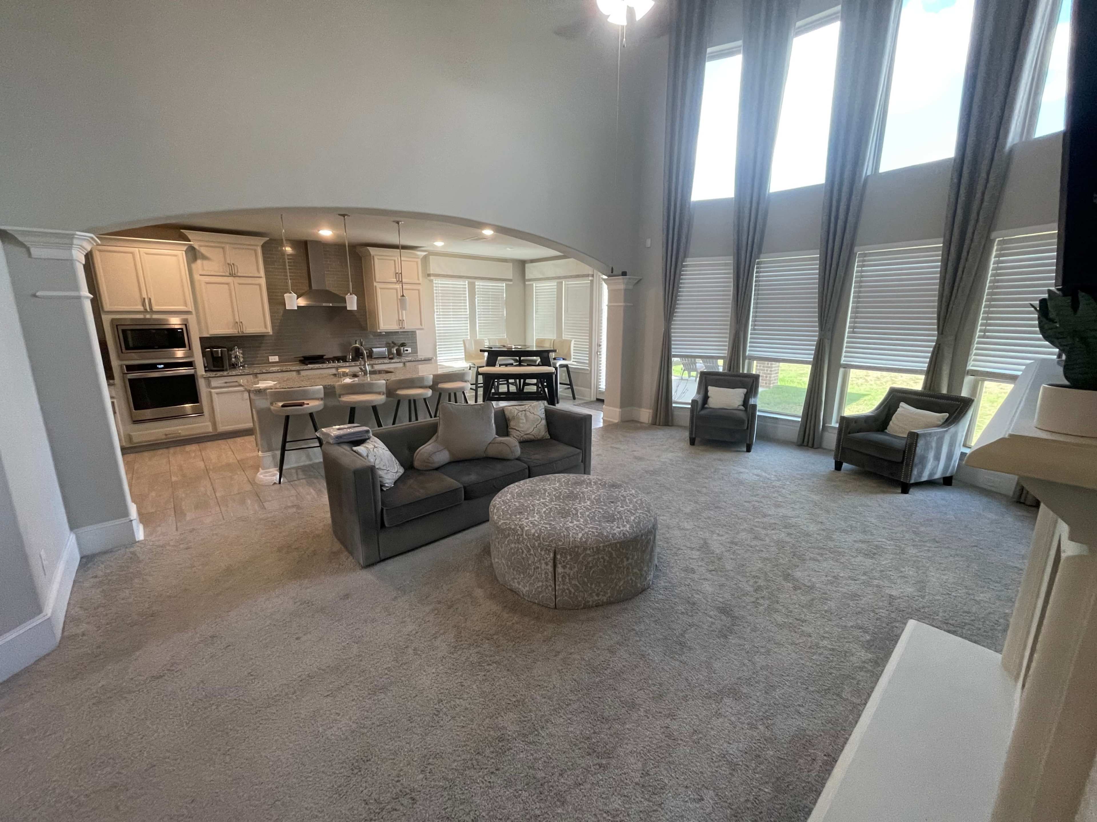 The image shows a spacious living room with a gray carpet, modern furnishings, and large windows allowing natural light to enter.