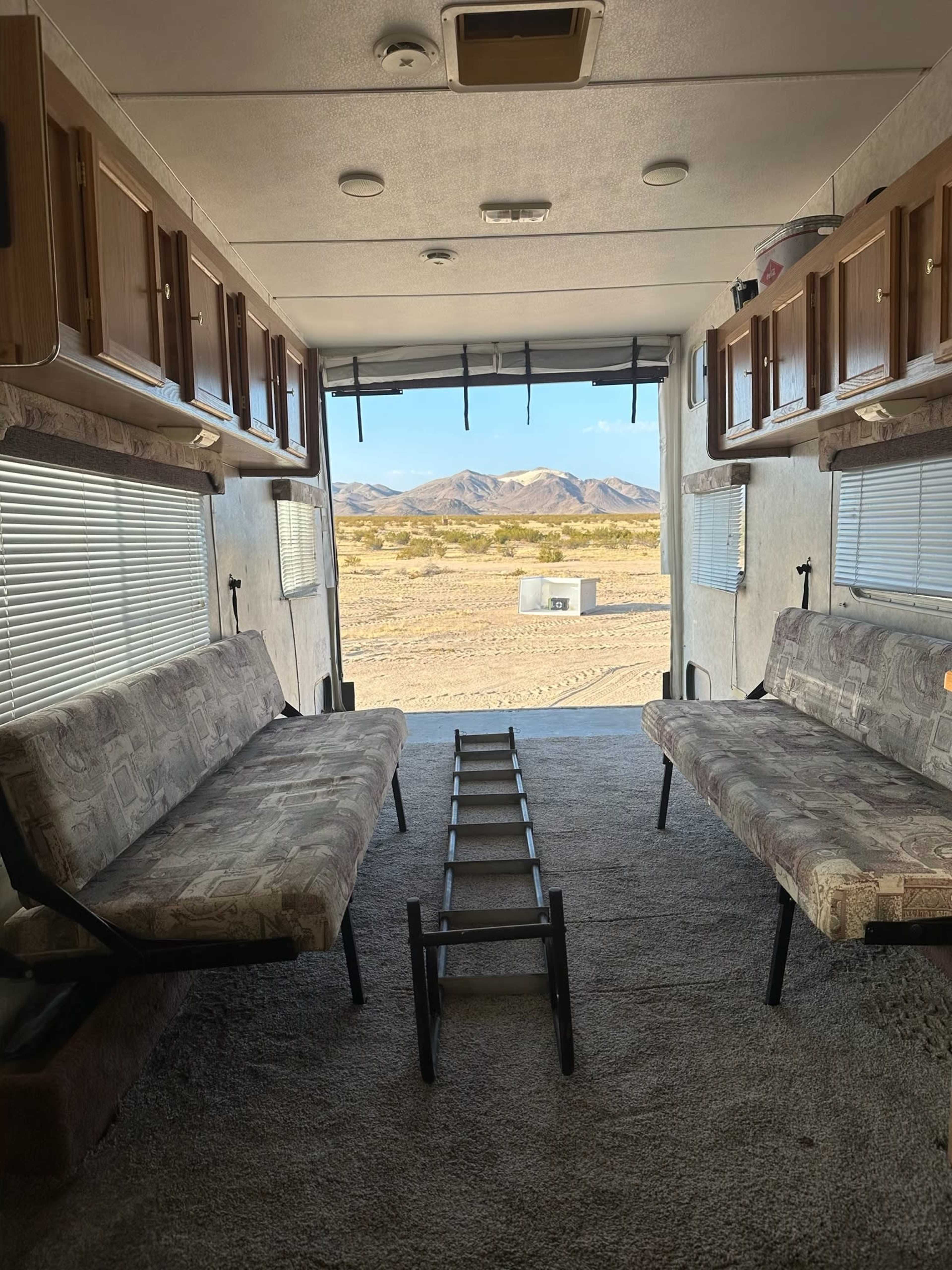 The interior of an RV shows two benches facing each other with a view of a desert landscape through an open rear door.