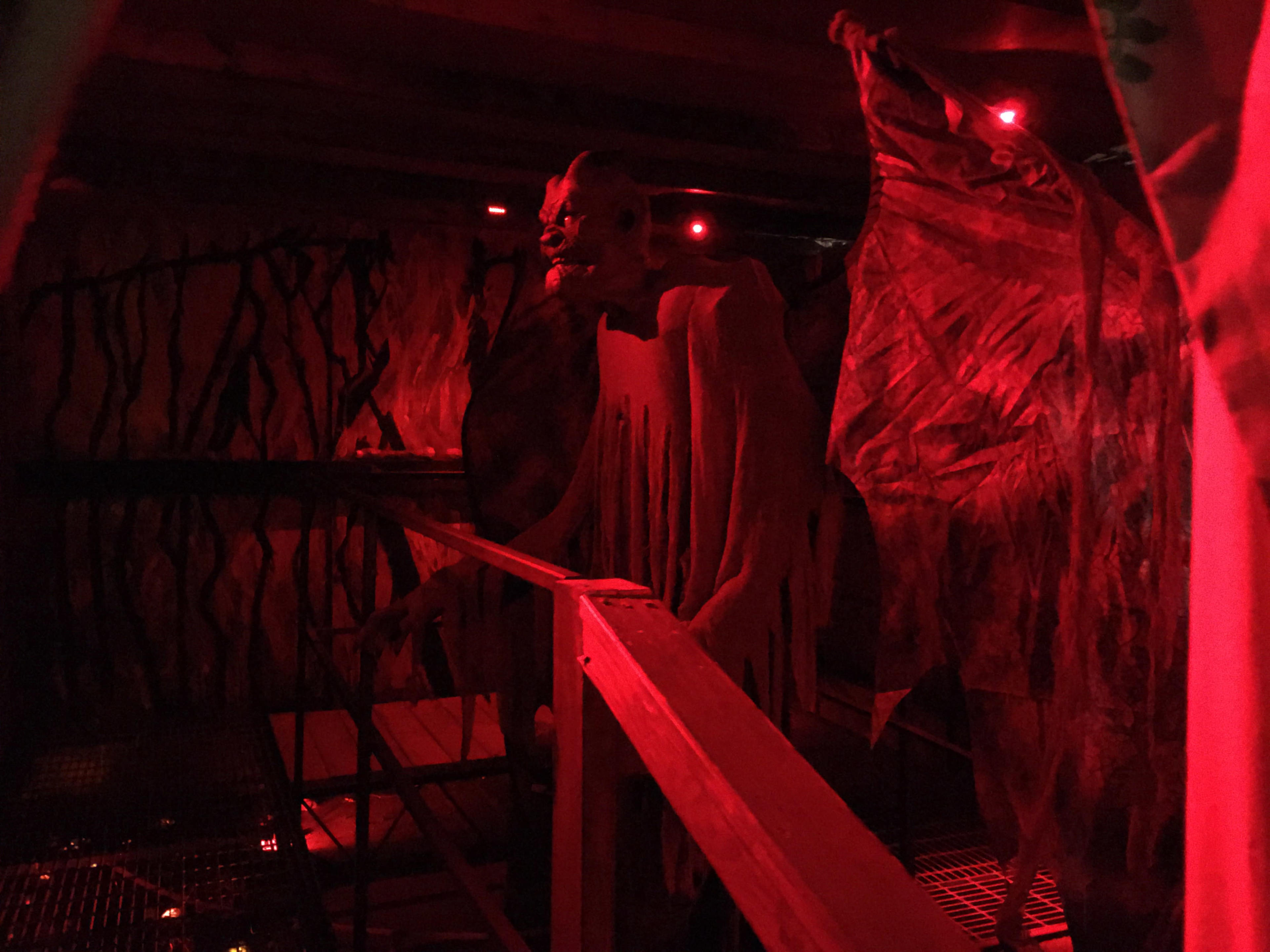 The dimly lit scene features eerie figures draped in tattered fabric against a red backdrop, with a wooden walkway in the foreground.
