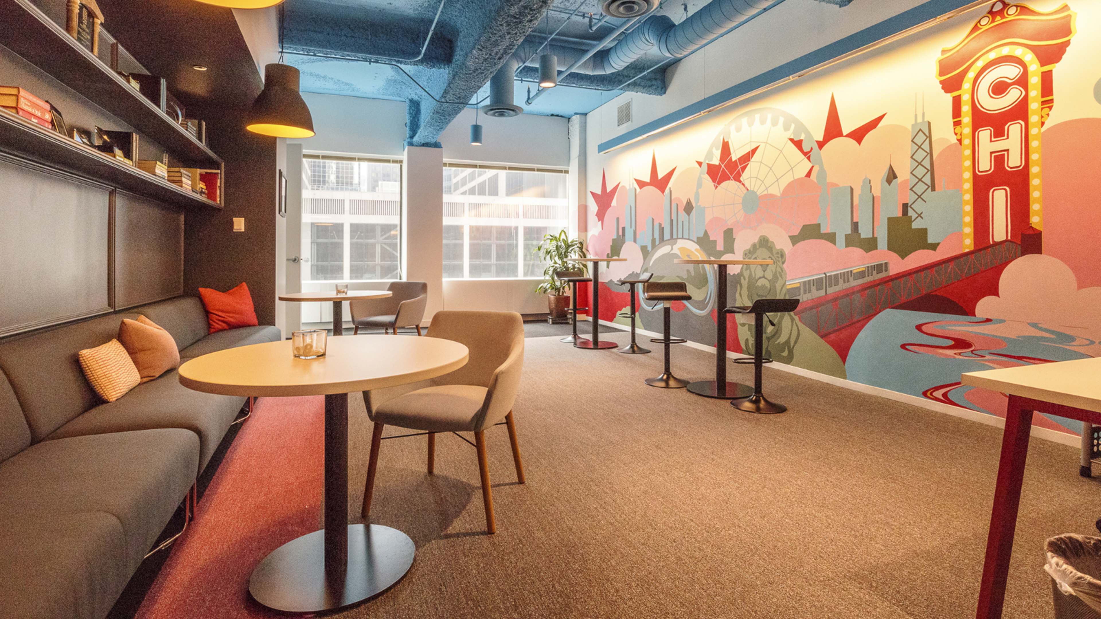A modern workspace with a gray lounge area, round tables, and a colorful mural featuring a bridge and skyline.