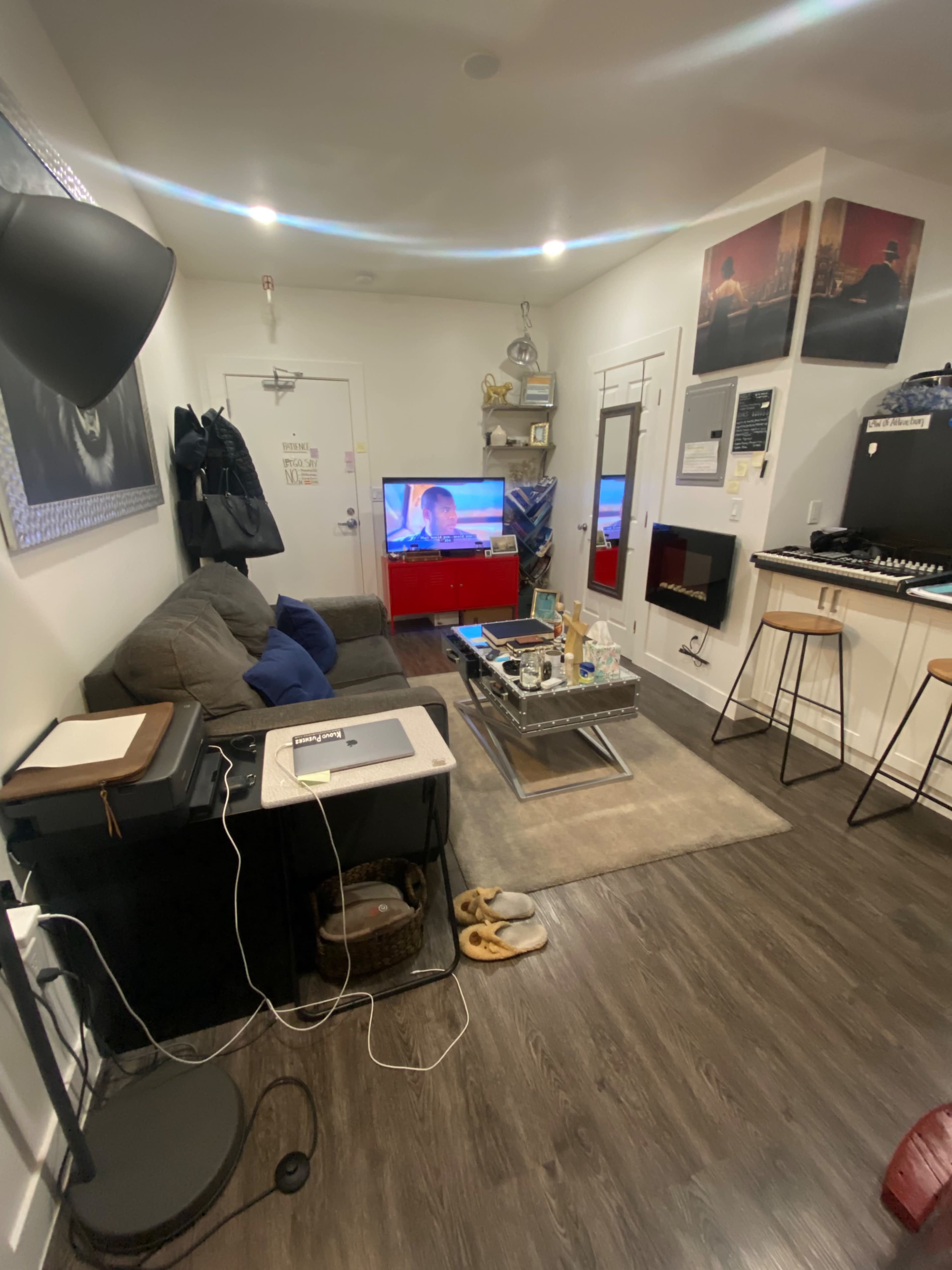 The living room features a couch, a television on a red stand, a coffee table with electronics, and a set of high stools next to a counter.