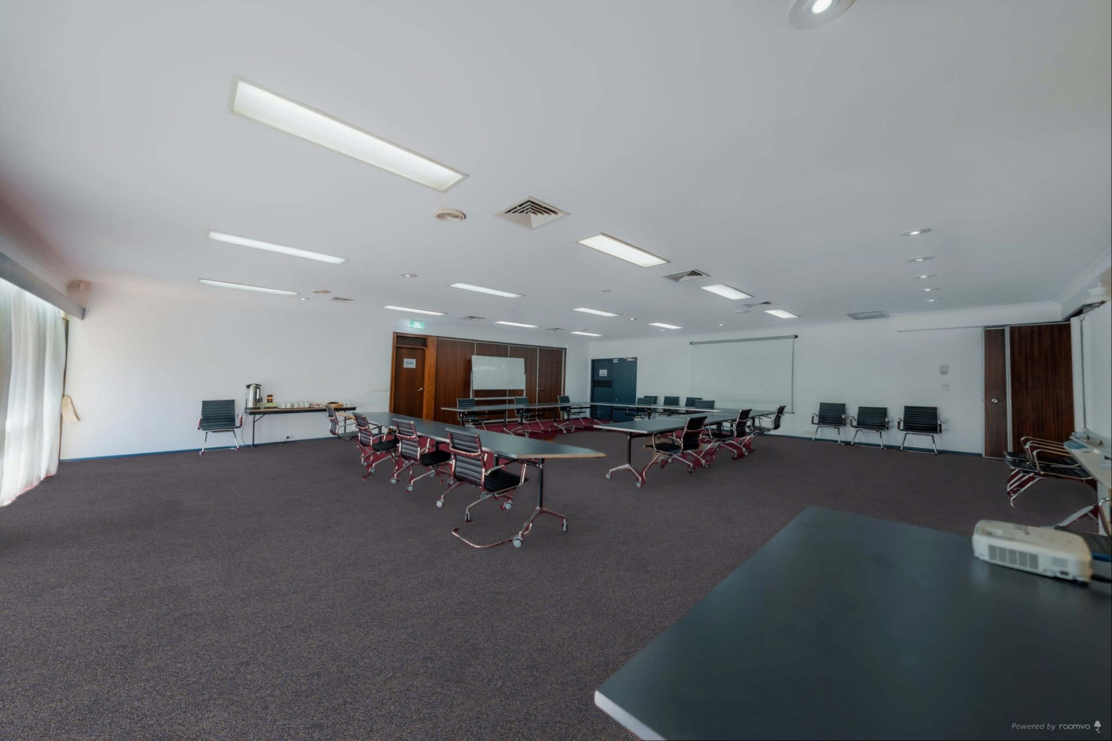 The image shows a large, empty meeting room with rows of chairs arranged around a central rectangular table and large windows letting in natural light.