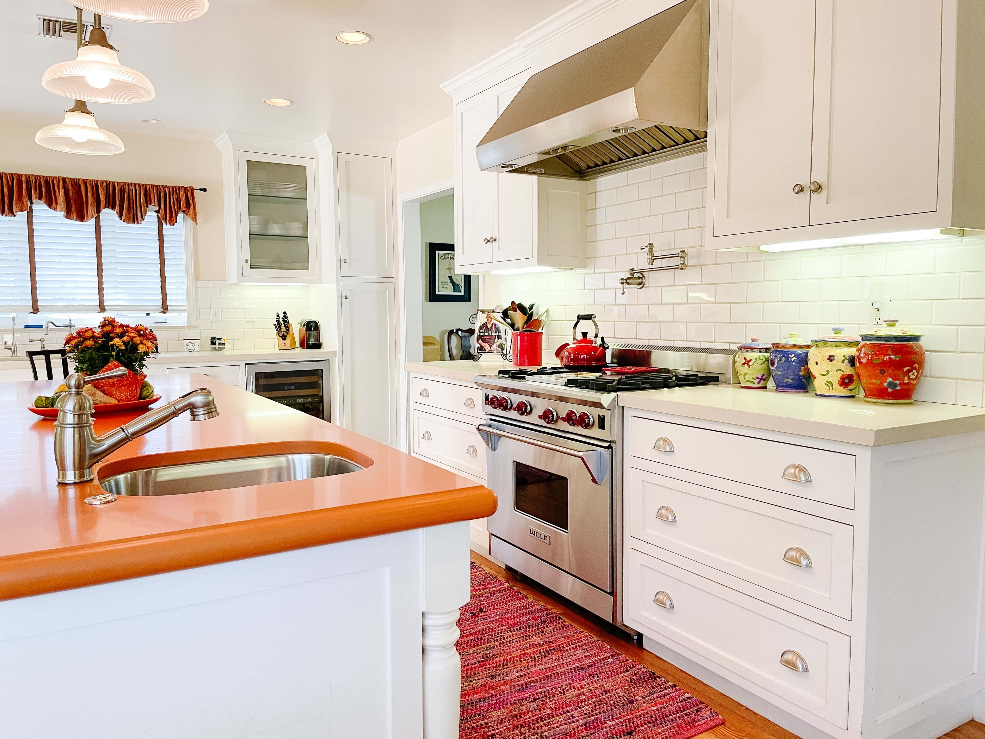 The kitchen features white cabinetry, a stainless steel range hood, and a vibrant orange countertop with a sink, complemented by colorful ceramic canisters and an island with a sink.