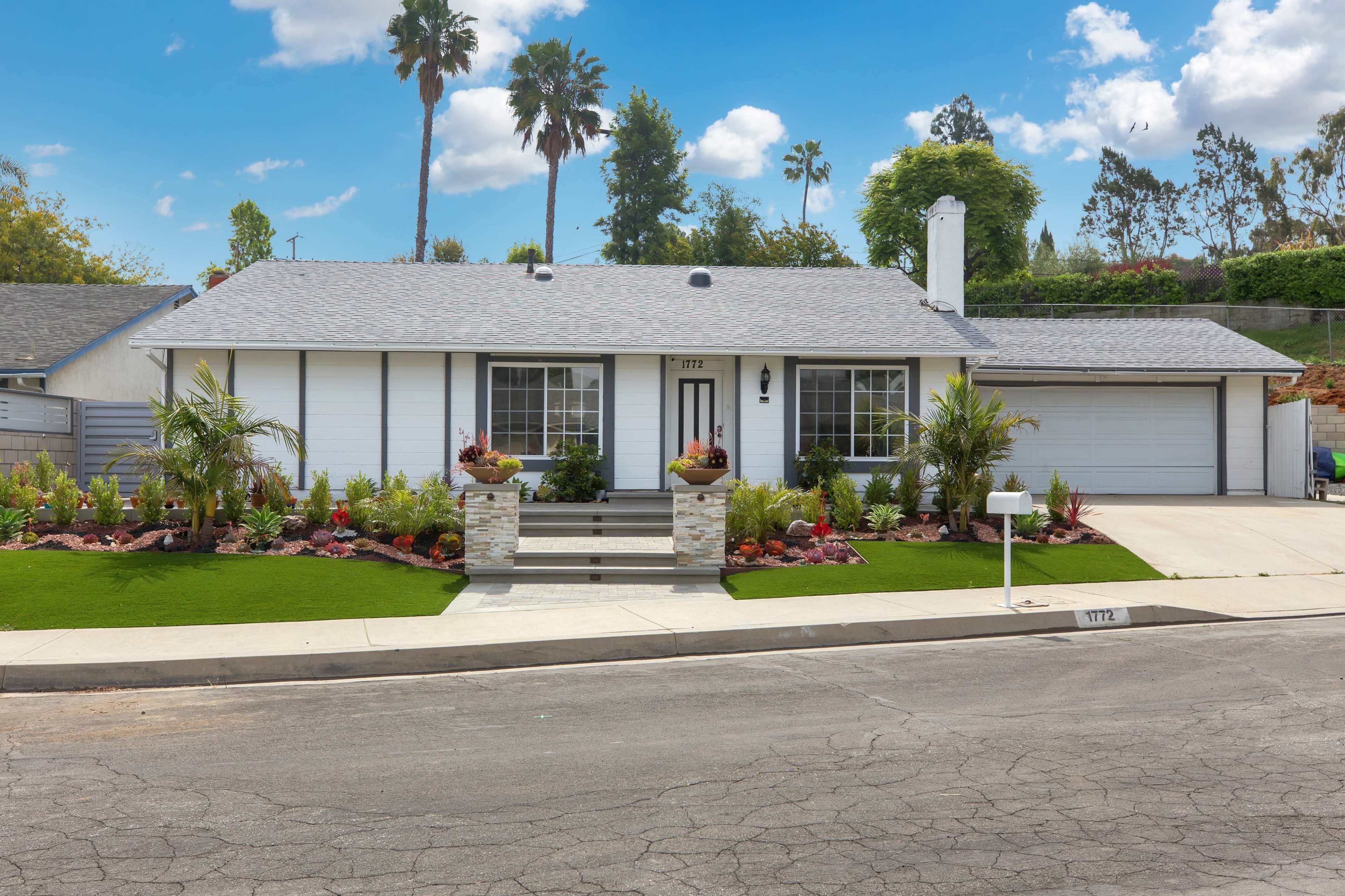 A single-story house with a landscaped front yard, featuring a stone pathway, flower beds, and a driveway.