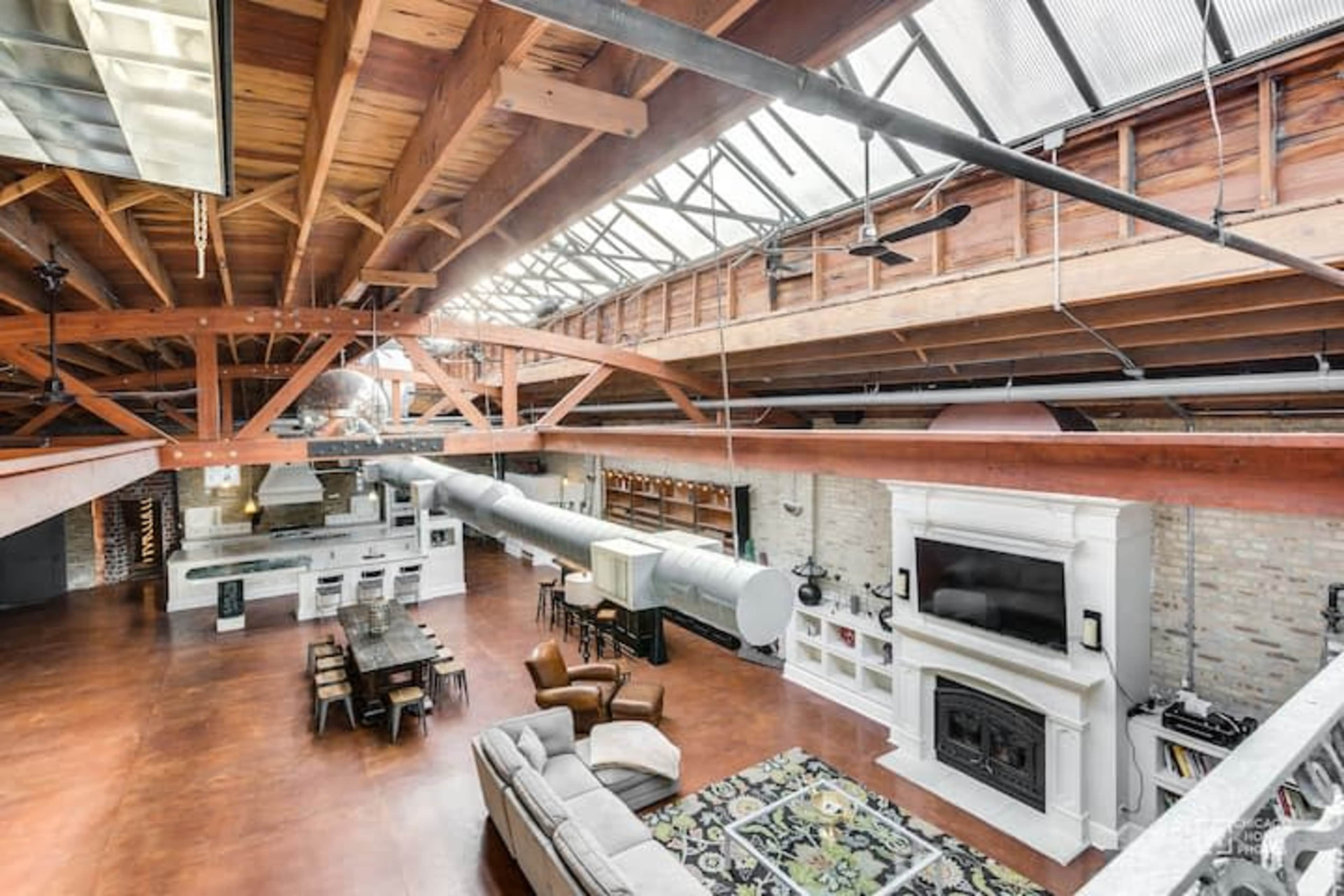 A spacious, loft-style living area with exposed wooden beams, large windows, and an open kitchen adjacent to a dining table and seating area.