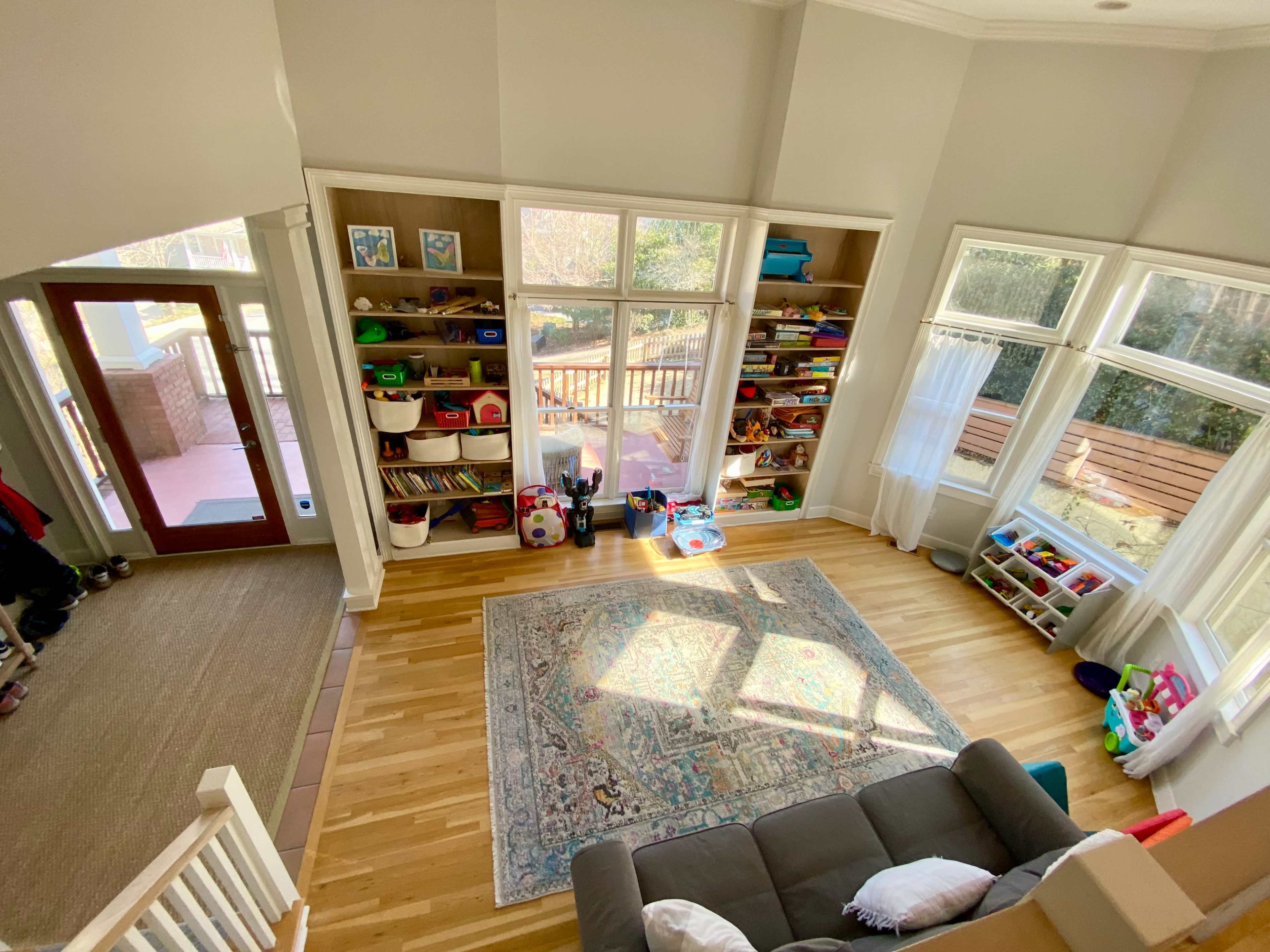 The image shows a bright, spacious room featuring a play area with shelves of toys, a large rug, and large windows overlooking a deck outside.