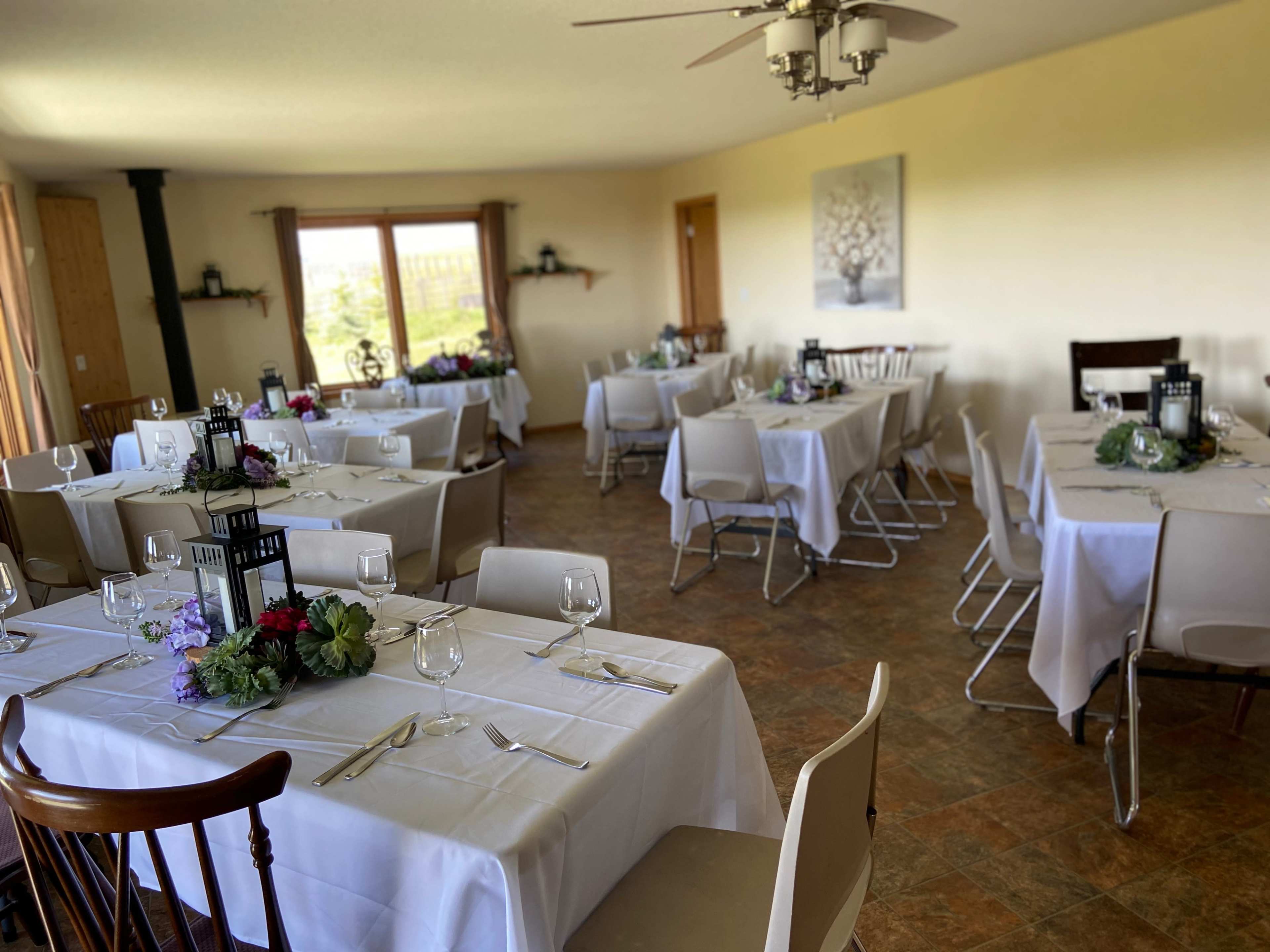 The image shows a dining room set for a meal, featuring multiple tables covered with white tablecloths and decorated with centerpieces, arranged in a spacious area with large windows.