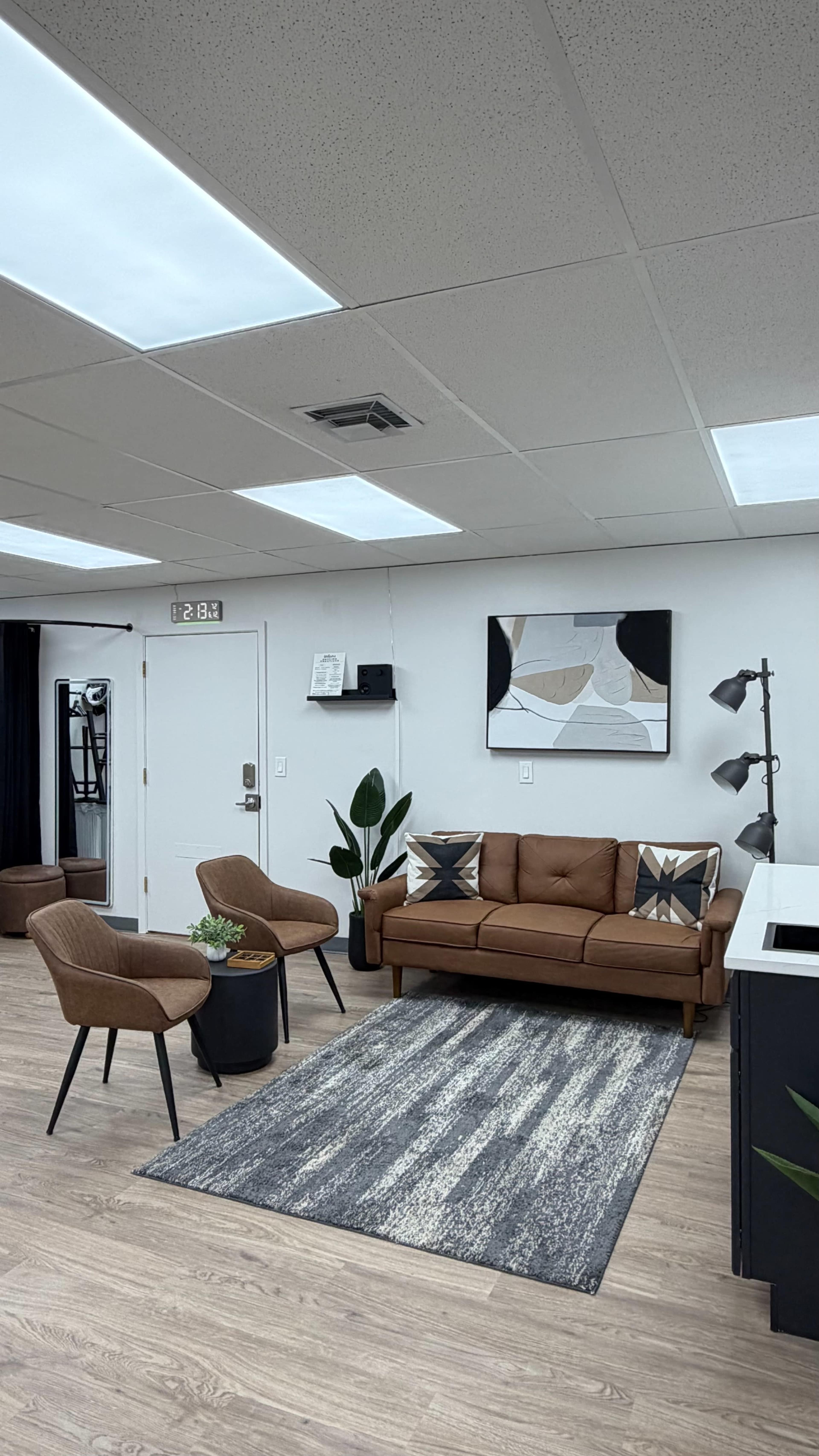 The image shows a modern indoor waiting area featuring a brown sofa, two chairs, a rug, and indoor plants, with ambient lighting from ceiling panels.