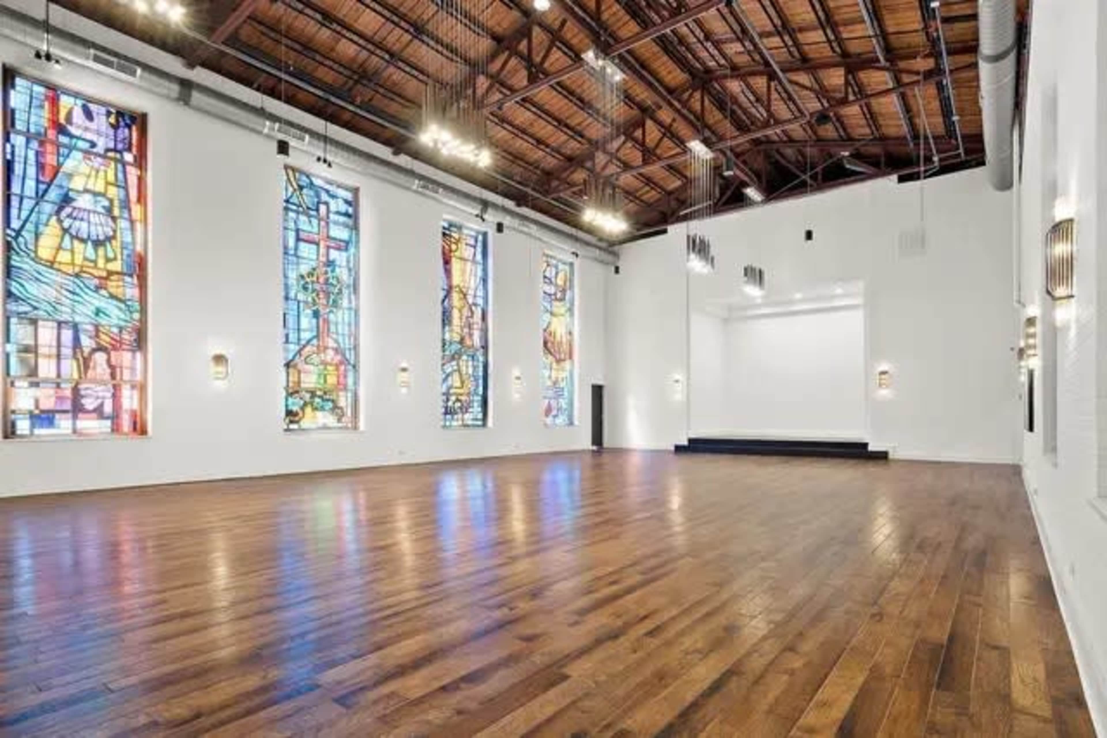 The image shows a spacious indoor venue with wooden flooring, featuring large stained glass windows on the walls and a raised stage area at the front.