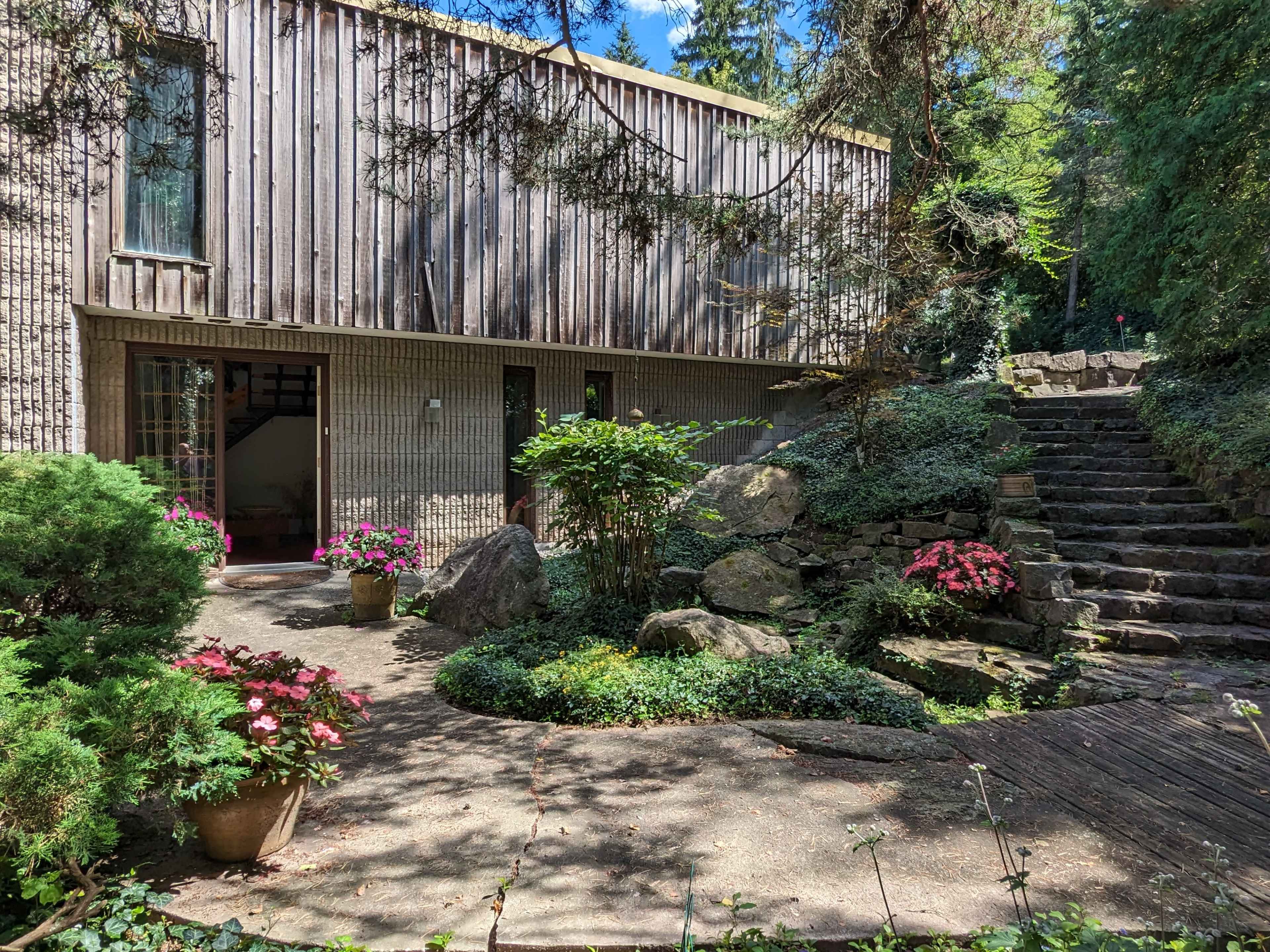 The image shows a modern wooden house surrounded by lush greenery and a stone pathway leading to a set of steps.