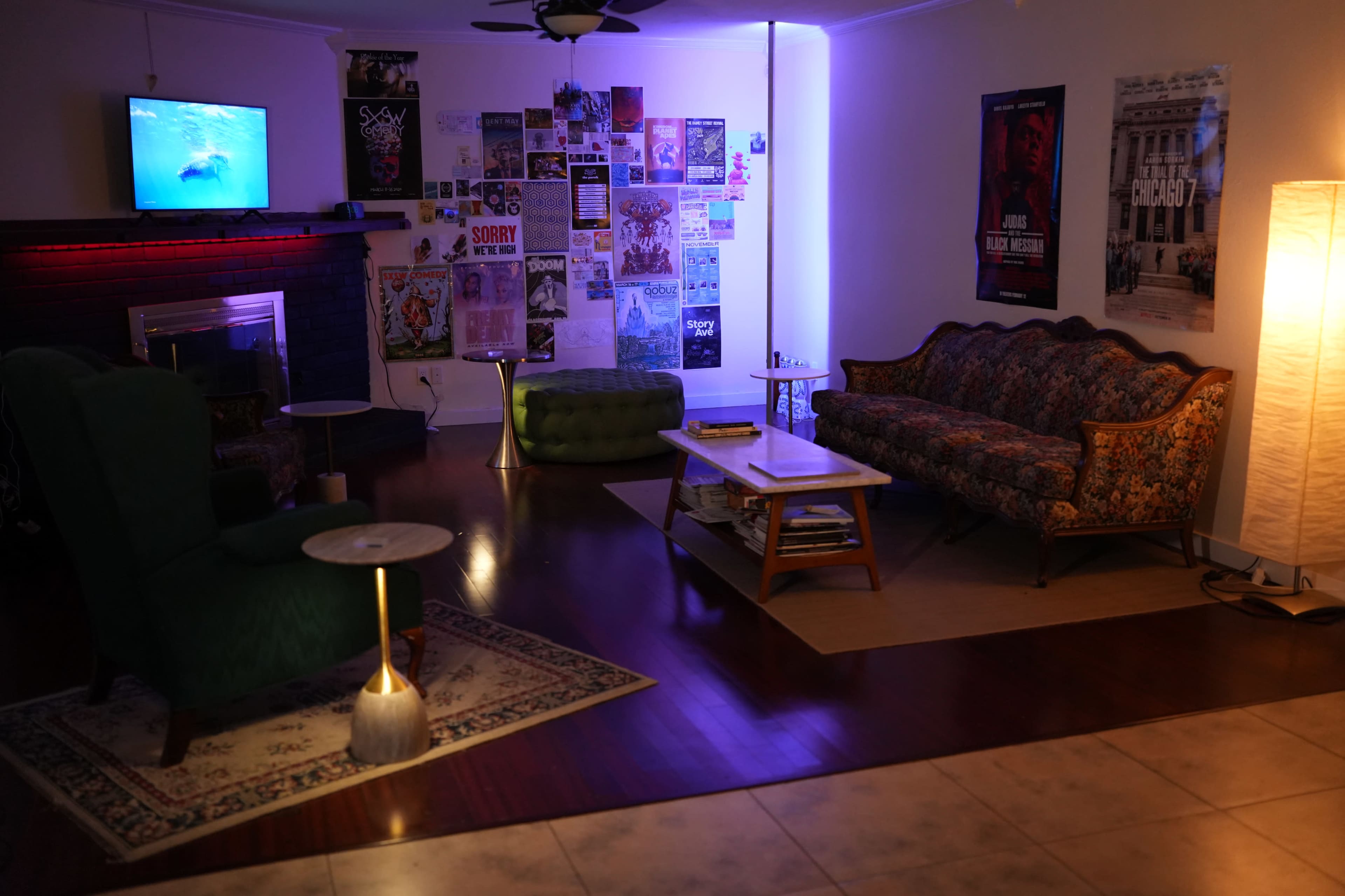 The room features a cozy seating area with a floral sofa, two armchairs, a coffee table, and a television mounted on the wall, surrounded by colorful posters and ambient lighting.