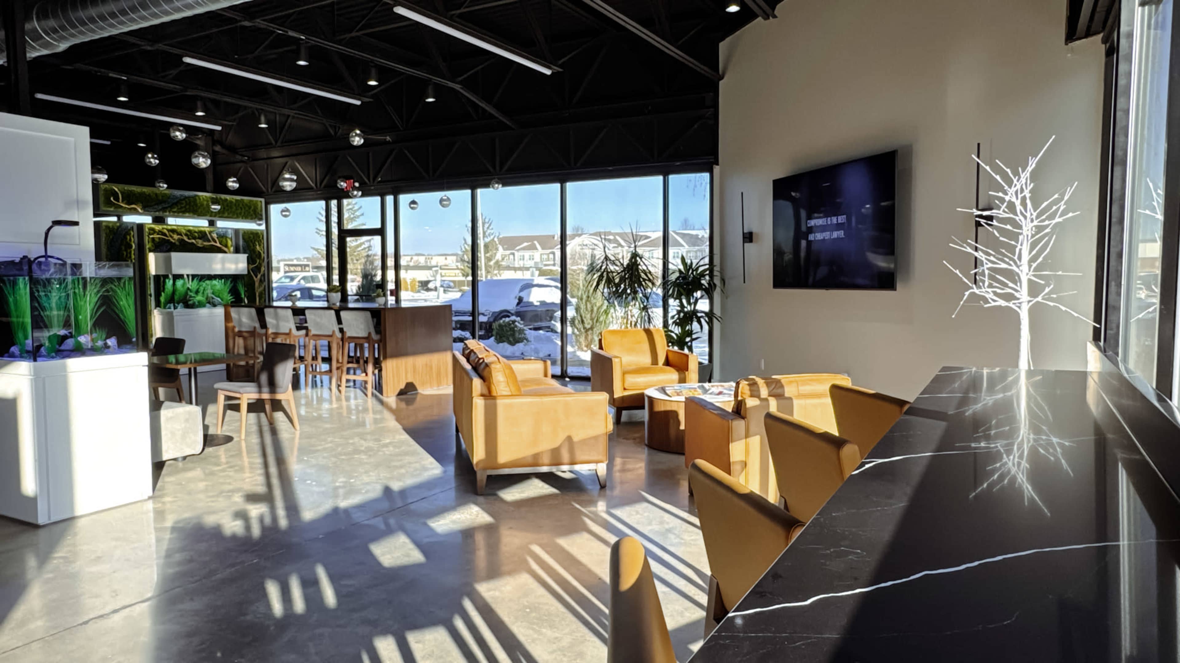 The image shows a modern interior space with large windows, featuring a lounge area with brown leather chairs, a bar table, and a sleek black counter.